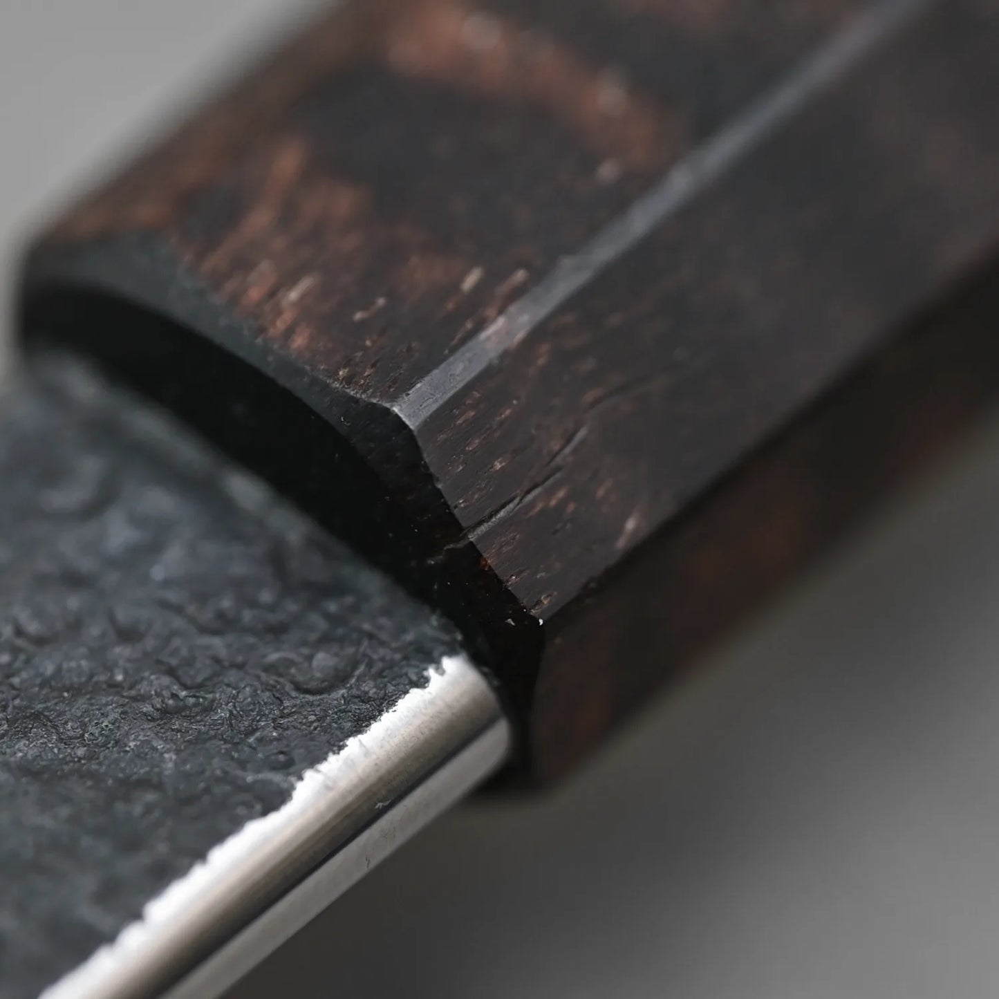 Close-up of the Krichbaum kurouchi wrought iron clad Apex Ultra gyuto 215x55, showing its dark, textured wood handle and hammered black blade finish in detail where the materials meet on this finely refinished knife.
