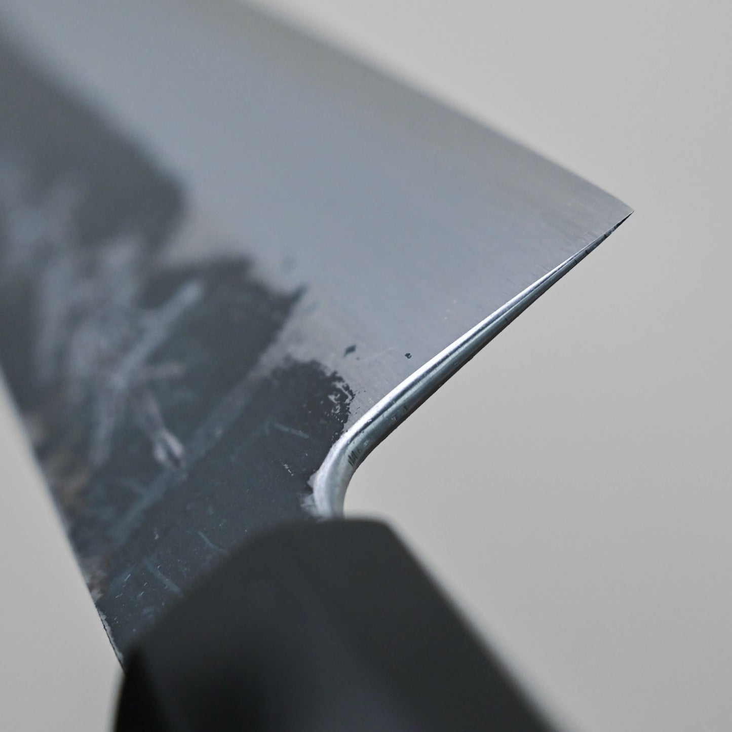 Close-up of the sharp edge and tip of the Yoshikazu Tanaka Ittosai Kotetsu Gokujo kurouchi shirogami#1 gyuto 240mm, featuring a Shirogami #1 steel gray blade and a black handle, set against a plain light background.