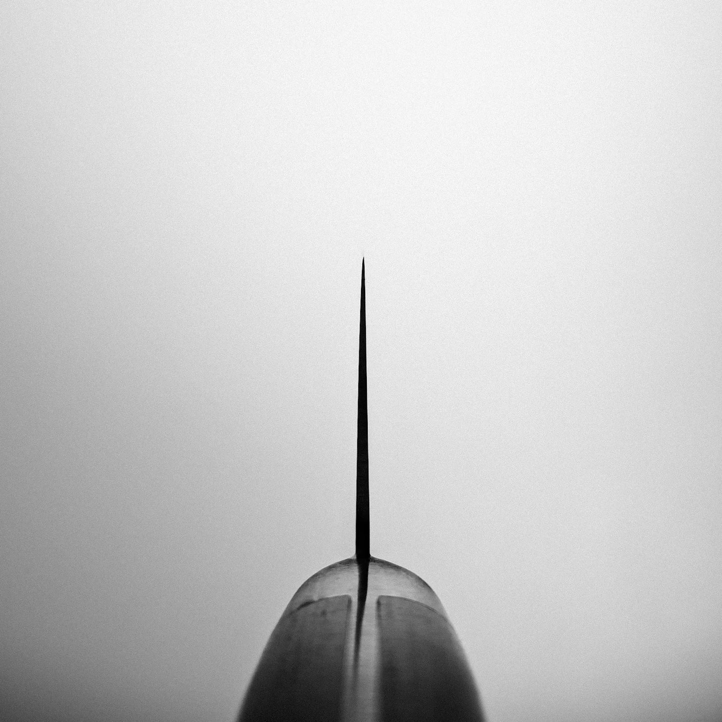 Close-up, black-and-white shot of the Misono Molybdenum gyuto 240mm knife blade tip, crafted from AUS8 stainless steel by Misono, pointing upward against a plain light background and centered in the frame.