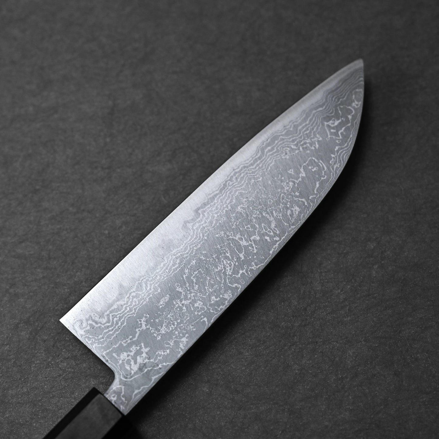 A close-up of the Hatsukokoro Damascus Aogami#2 Santoku 165mm knife shows its distinctive Damascus steel blade against a dark, textured background.