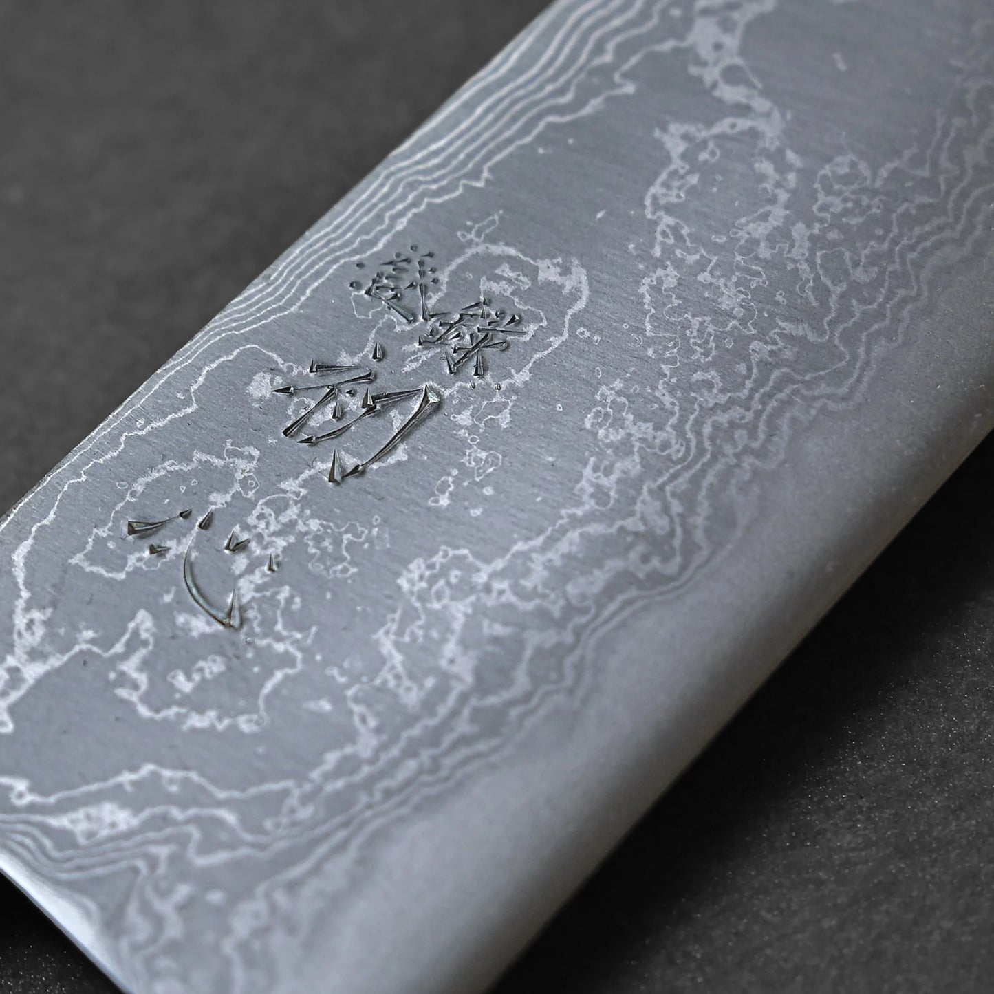 Close-up of the Hatsukokoro damascus aogami#2 santoku 165mm knife, showcasing its wave-patterned Damascus steel and engraved Japanese kanji, set against a dark background.