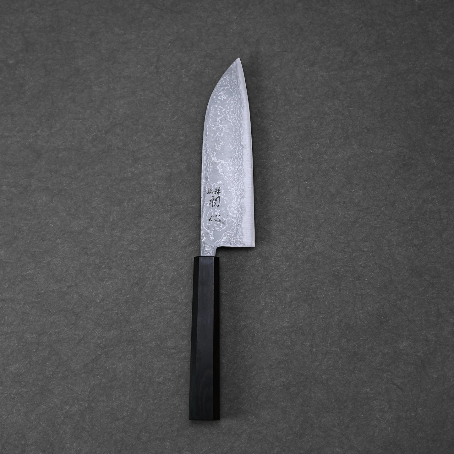 The Hatsukokoro damascus aogami#2 santoku 165mm knife, featuring a patterned blade and black rectangular handle, rests on a dark grey textured surface.