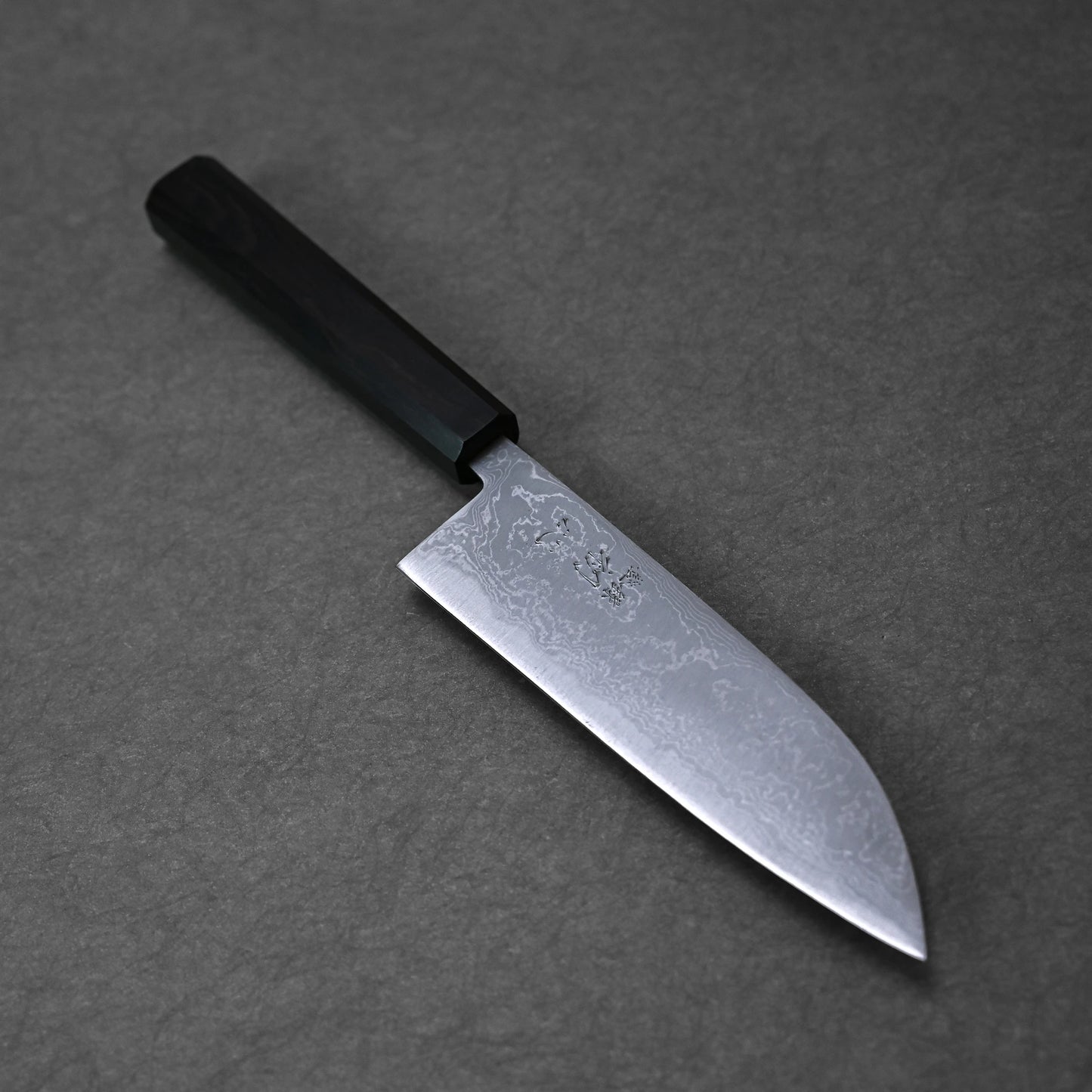 The Hatsukokoro damascus aogami#2 santoku 165mm, crafted by Hatsukokoro, features a dark handle and elegant patterned steel blade, resting on a grey textured surface.