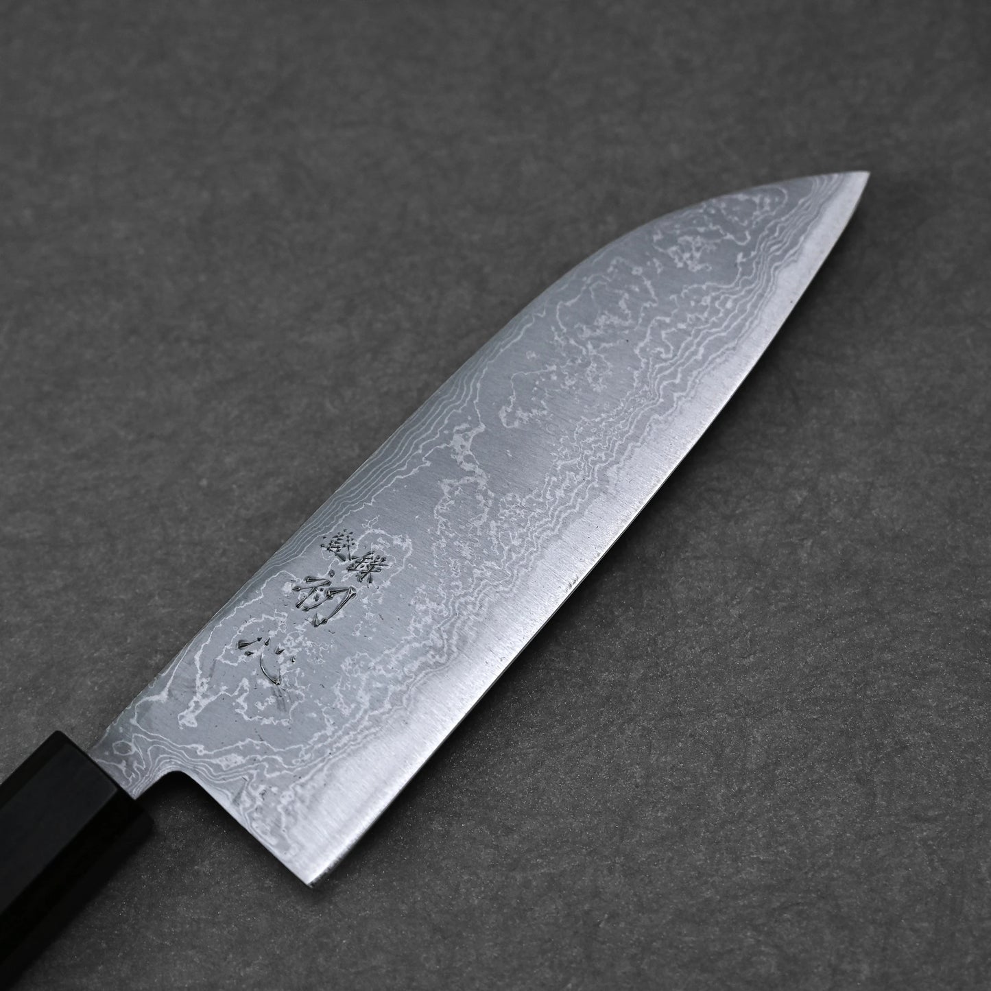 The Hatsukokoro damascus aogami#2 santoku 165mm features a patterned Damascus steel blade with engraved Japanese characters and a black handle, shown resting on a dark grey textured surface.