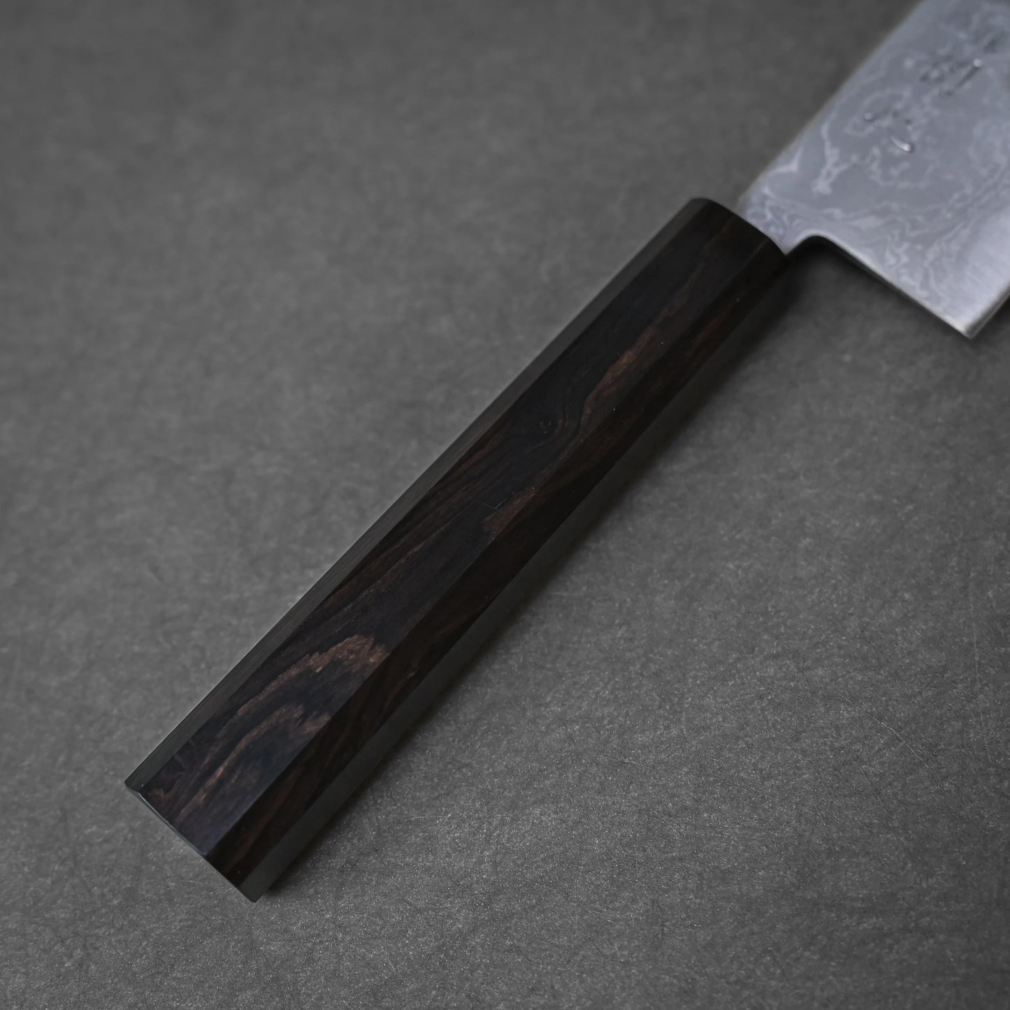 A close-up of the Hatsukokoro damascus aogami#2 santoku 165mm shows its dark wooden handle with subtle grain and part of its silver blade, set against a textured grey background.