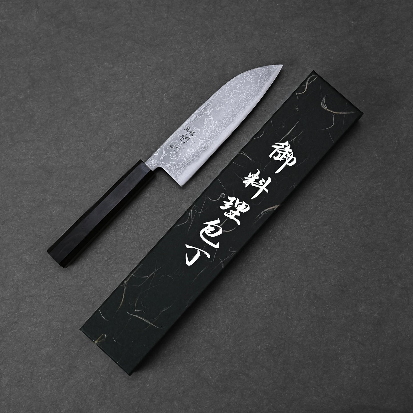 The Hatsukokoro damascus aogami#2 santoku 165mm, made by Hatsukokoro with Aogami #2 carbon steel, features a dark handle and is shown beside its black box with white Japanese characters on a dark textured background.