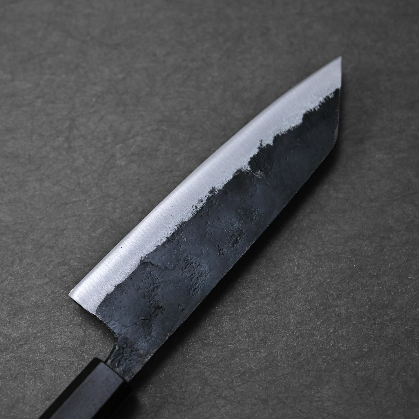 A close-up of the Hatsukokoro Kurouchi Aogami#2 Bunka 165mm knife, handforged in Tosa, Japan, featuring a dark textured kurouchi finish and polished edge, by Hatsukokoro, displayed on a dark gray surface.