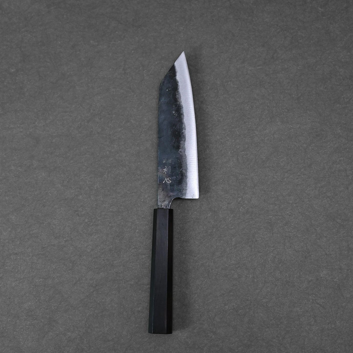 The Hatsukokoro kurouchi aogami#2 bunka 165mm, a Japanese chefs knife with a dark wooden handle and kurouchi-finished blade, rests on a gray textured surface; handforged in Tosa, Japan by Hatsukokoro.