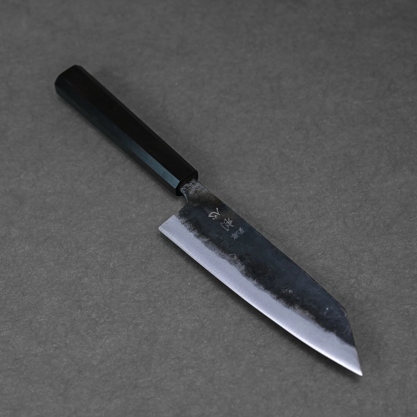 The Hatsukokoro kurouchi aogami#2 bunka 165mm, a Japanese kitchen knife with a black handle and rustic kurouchi finish, rests on gray textured surface. Handforged in Tosa Japan, it features a pointed tip and visible textured blade edge.