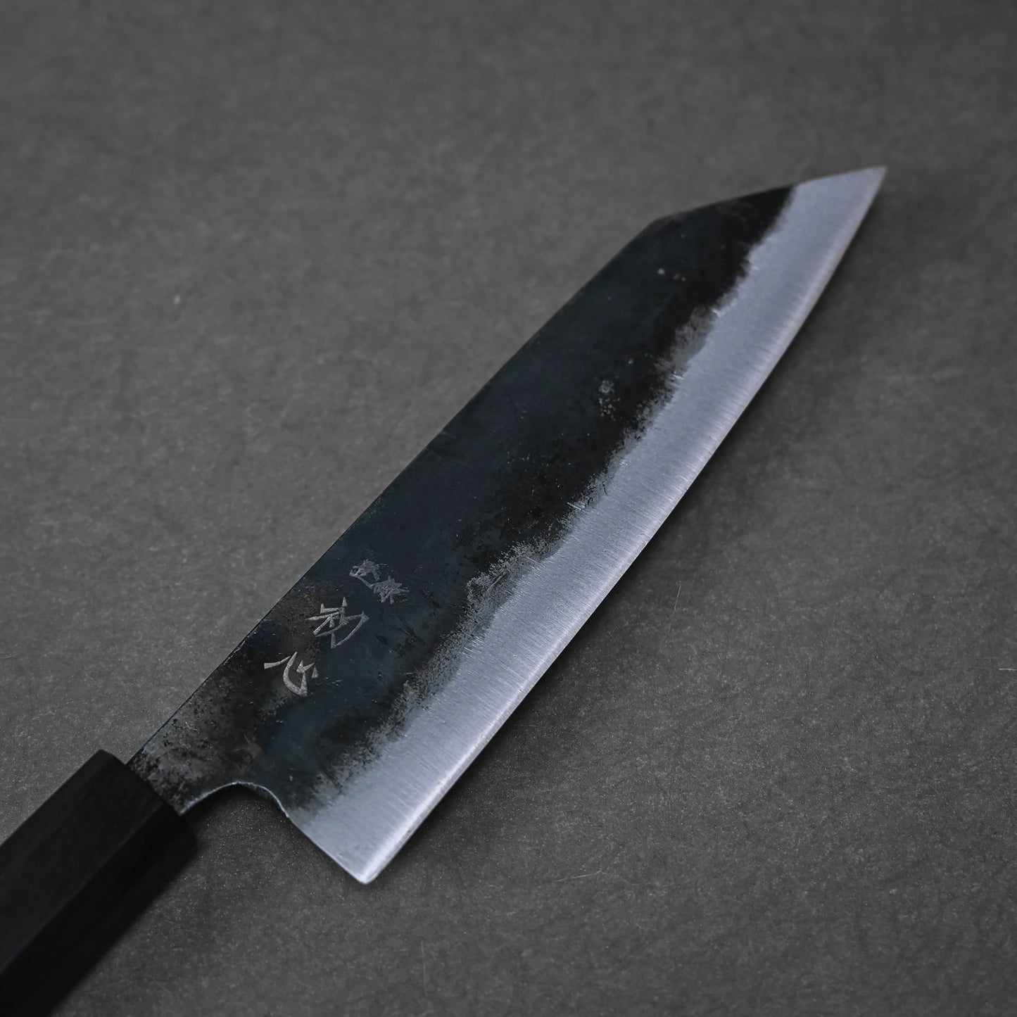 The Hatsukokoro kurouchi aogami#2 bunka 165mm, a Japanese kitchen knife by Hatsukokoro, features a handforged matte dark blade with kurouchi finish and engraved markings near its rectangular dark handle, shown resting on a gray surface.
