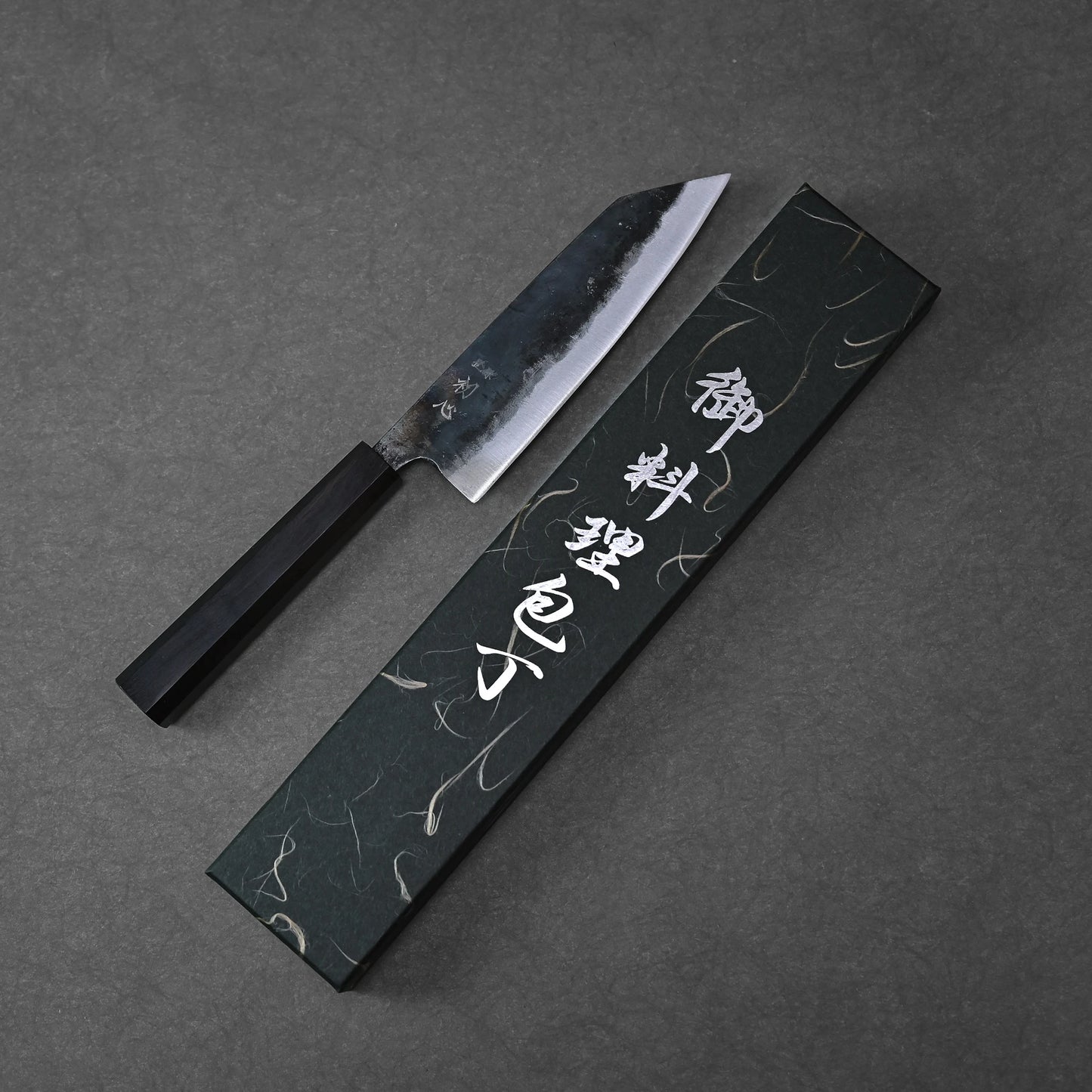The Hatsukokoro kurouchi aogami#2 bunka 165mm, a Japanese kitchen knife with a dark handle and kurouchi finish, is handforged by Hatsukokoro in Tosa Japan and displayed beside its black box with white Japanese characters on a gray surface.