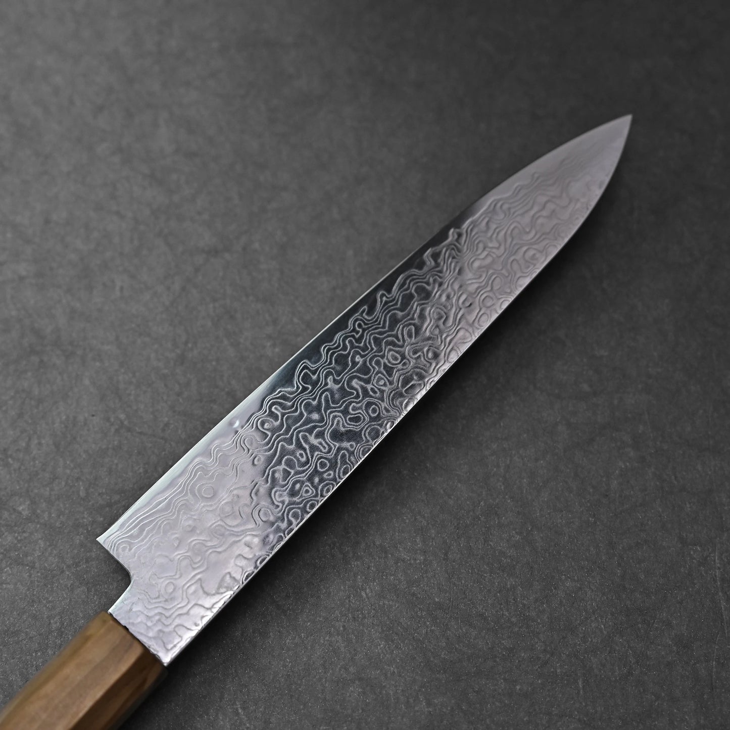 Reverse side of an SG2 damascus petty knife with olive wood handle; photographed to showcase the flowing damascus pattern across the blade, SG2 steel marking near the base, and the gentle curvature of the olive wood handle set against a dark background.