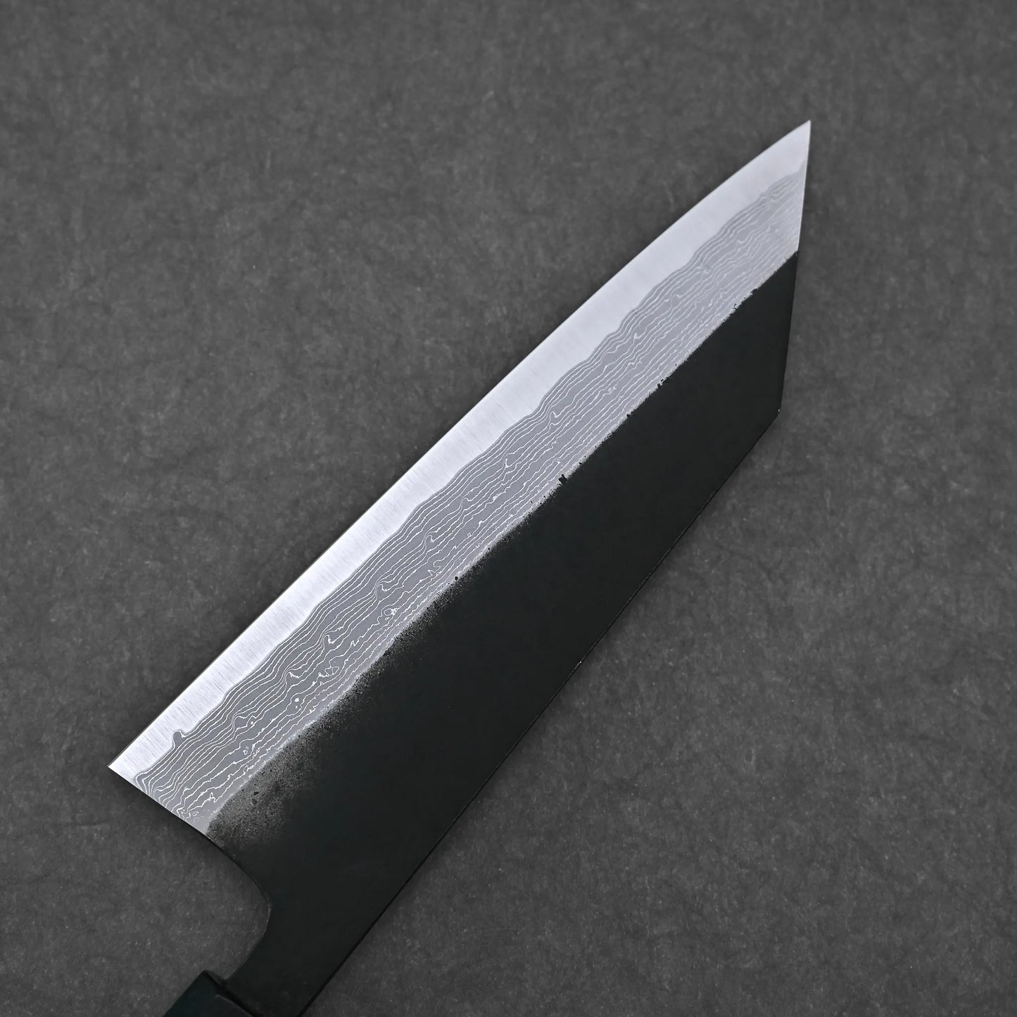 A close-up of the Hatsukokoro Shinkiro kurouchi damascus AS bunka 170mm knife blade, showcasing its wavy Damascus steel patterns on the polished edge against a dark gray textured background.