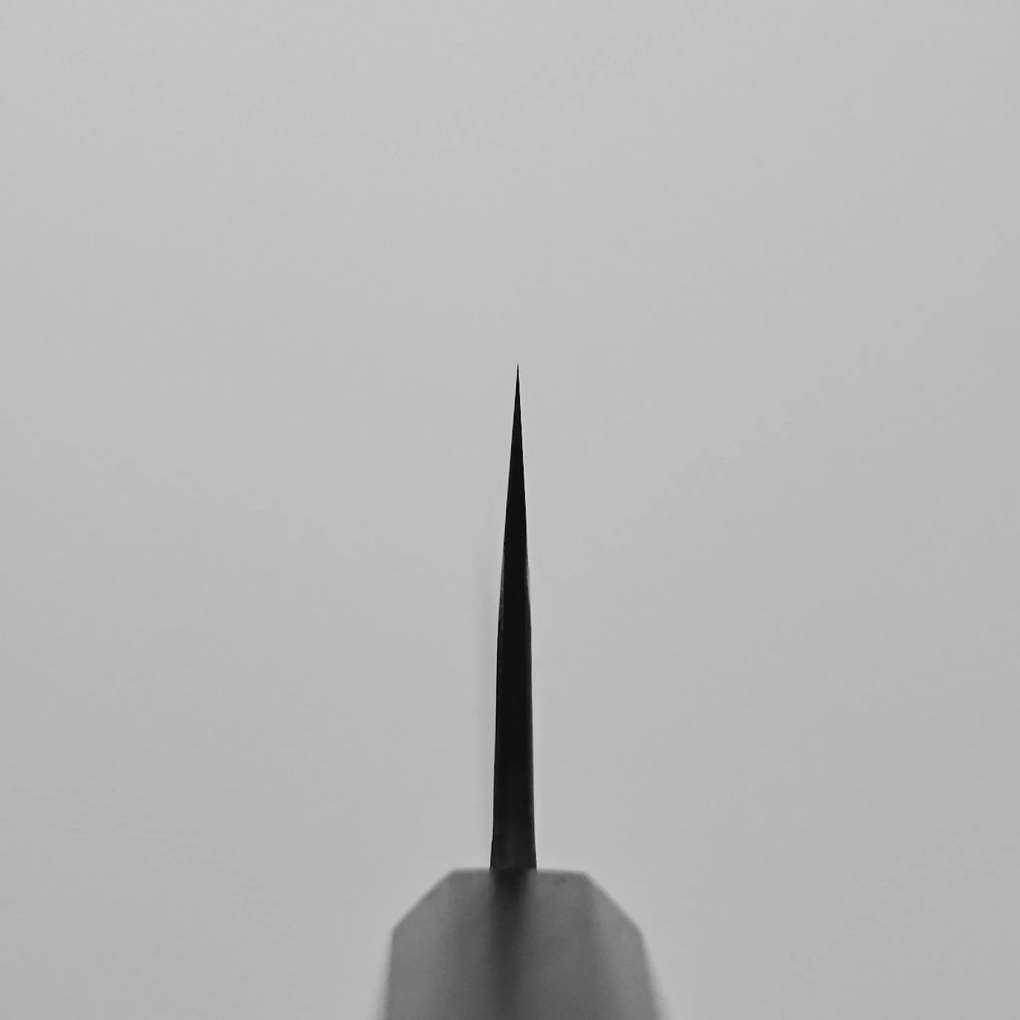 Close-up of the Hatsukokoro Shinkiro kurouchi damascus AS bunka 170mm (no logo) knife tip, shot from the front with a shallow depth of field; the thin, sharp blade stands out against a plain light gray background.