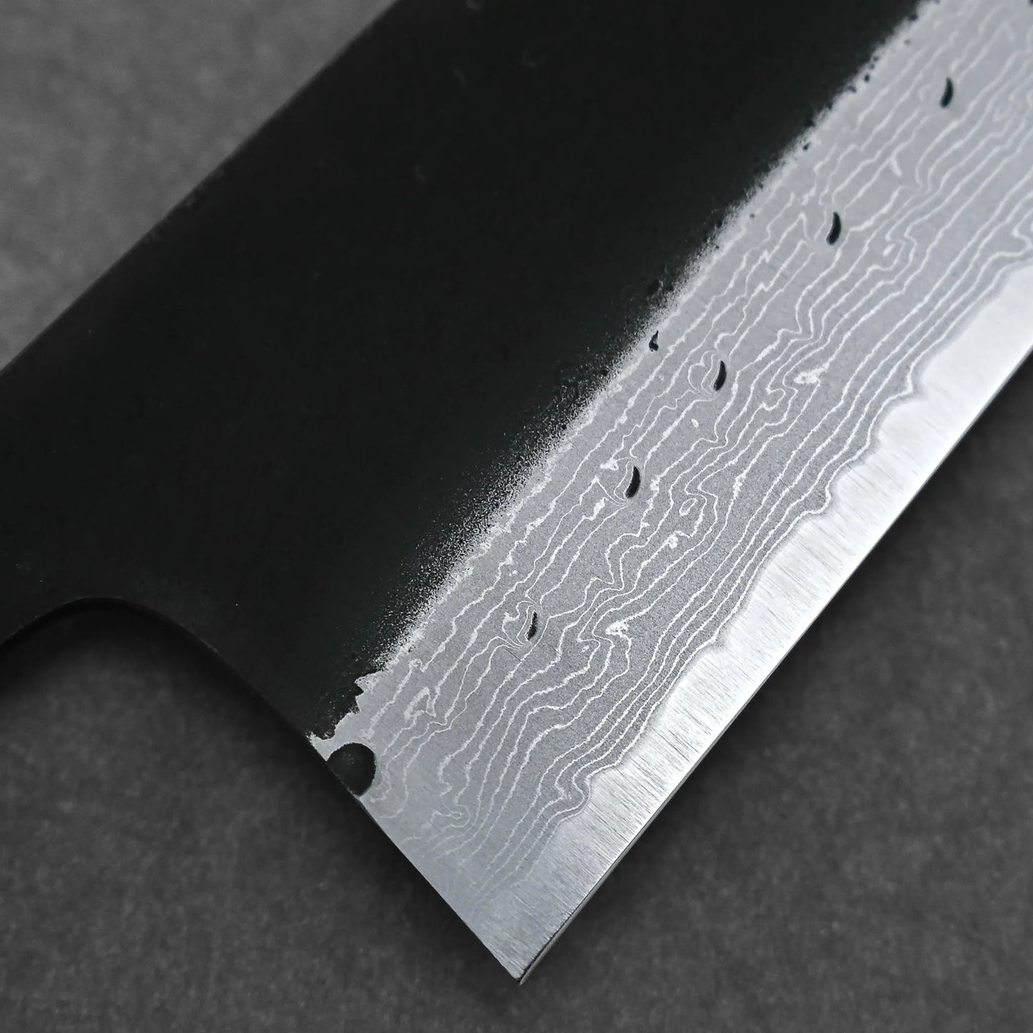 Close-up of the Hatsukokoro Shinkiro kurouchi damascus AS bunka 170mm (no logo), highlighting its wavy Damascus steel and matte black handle, set against a dark, textured background.