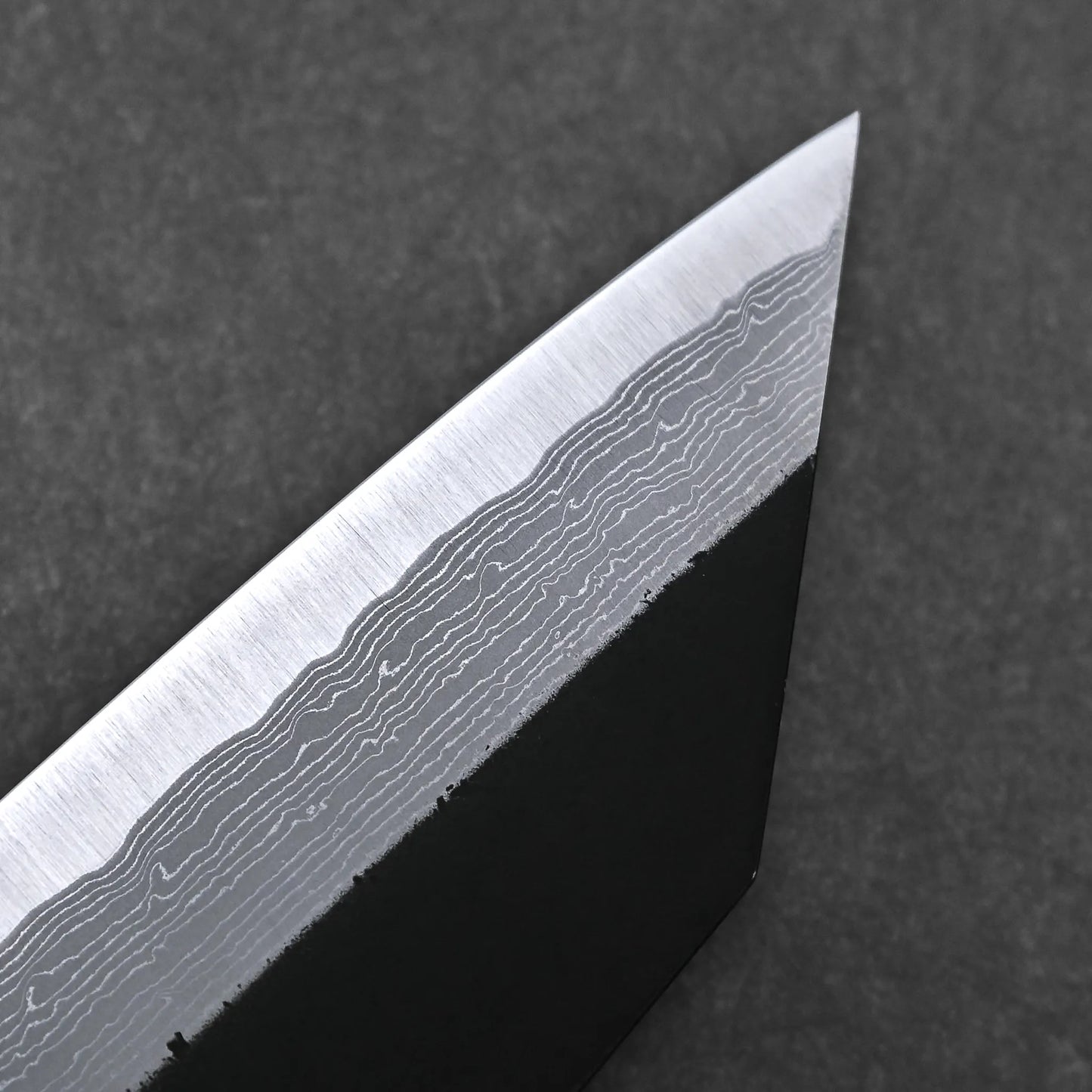 Close-up of the Hatsukokoro Shinkiro kurouchi damascus AS bunka 170mm blade, showcasing its wavy Damascus steel pattern and sharp pointed tip against a dark background.