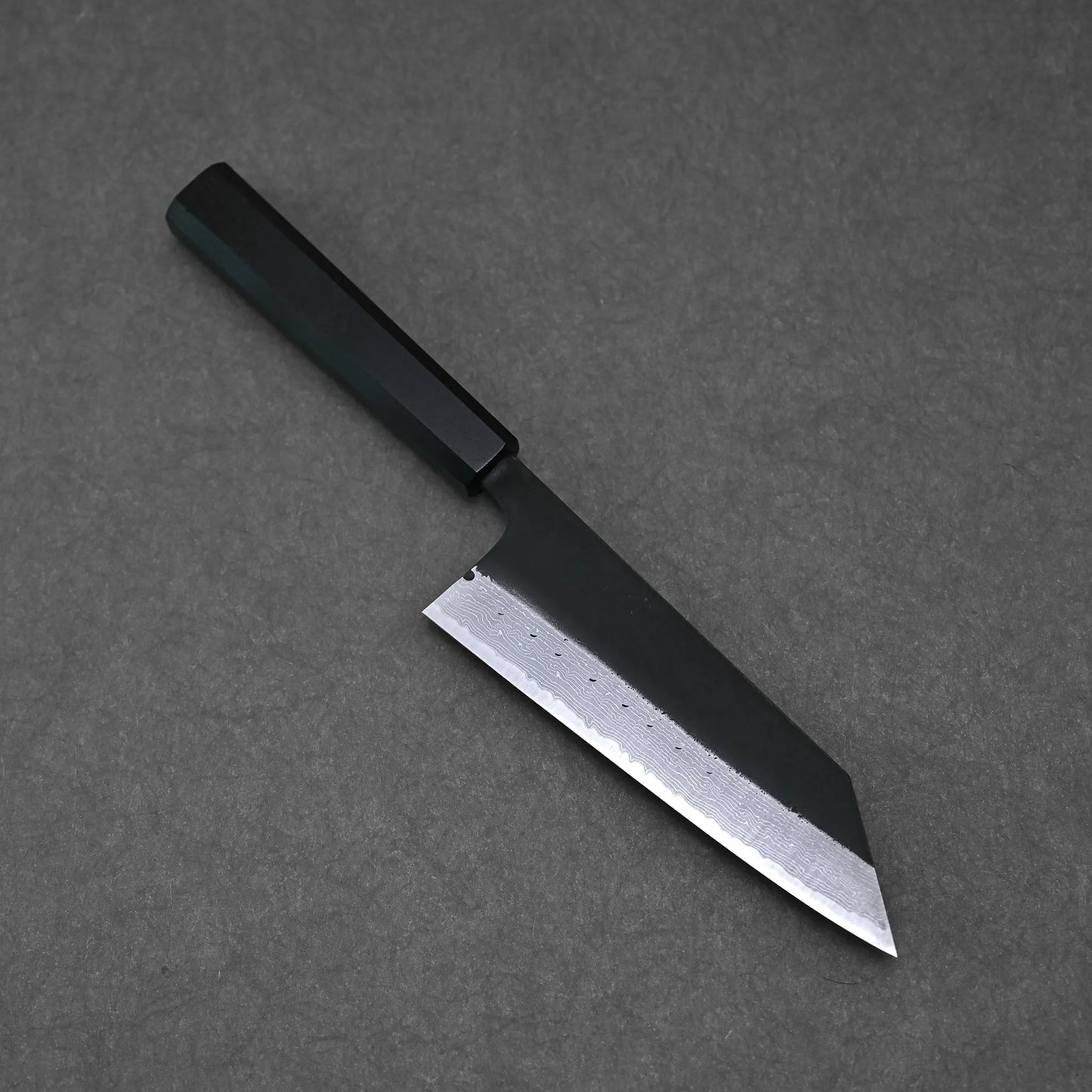 A Hatsukokoro Shinkiro kurouchi damascus AS bunka 170mm knife with a straight edge, pointed tip, and dark handle rests on a flat gray surface.