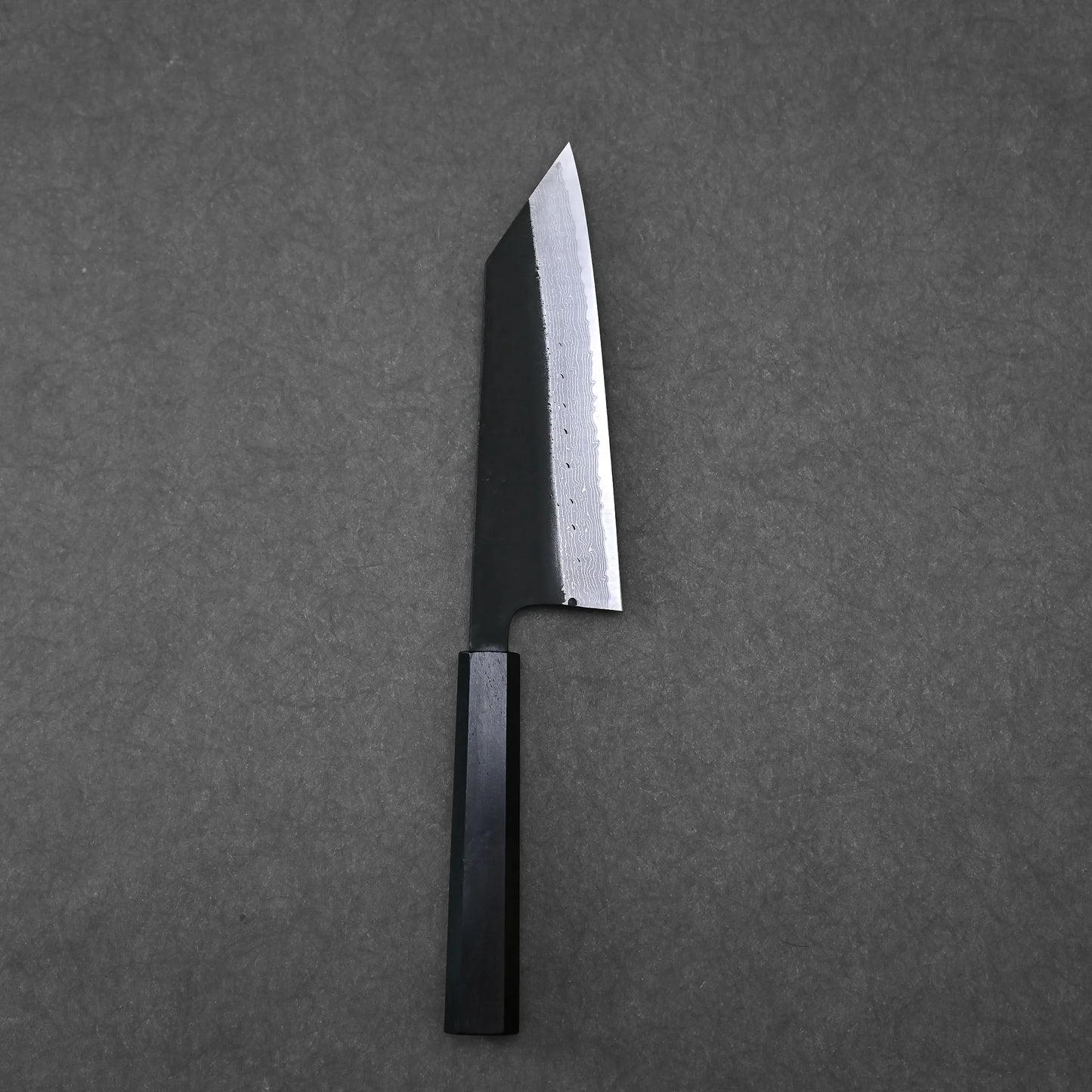 The Hatsukokoro Shinkiro kurouchi damascus AS bunka 170mm - no logo, a hand-forged Japanese knife with a dark handle and broad, angled blade, is displayed on a dark gray textured surface.