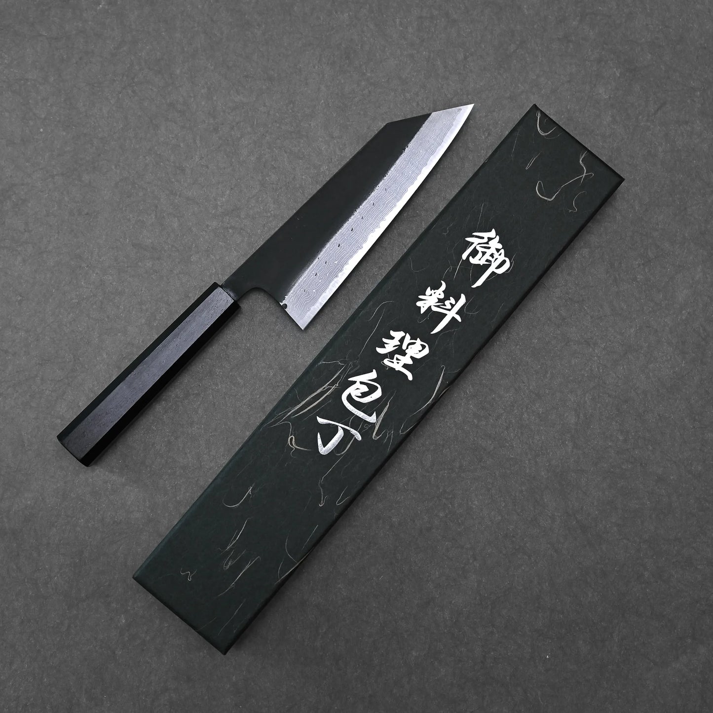 The Hatsukokoro Shinkiro kurouchi damascus AS bunka 170mm knife with a black handle lies beside a sleek black box on a dark, textured surface.