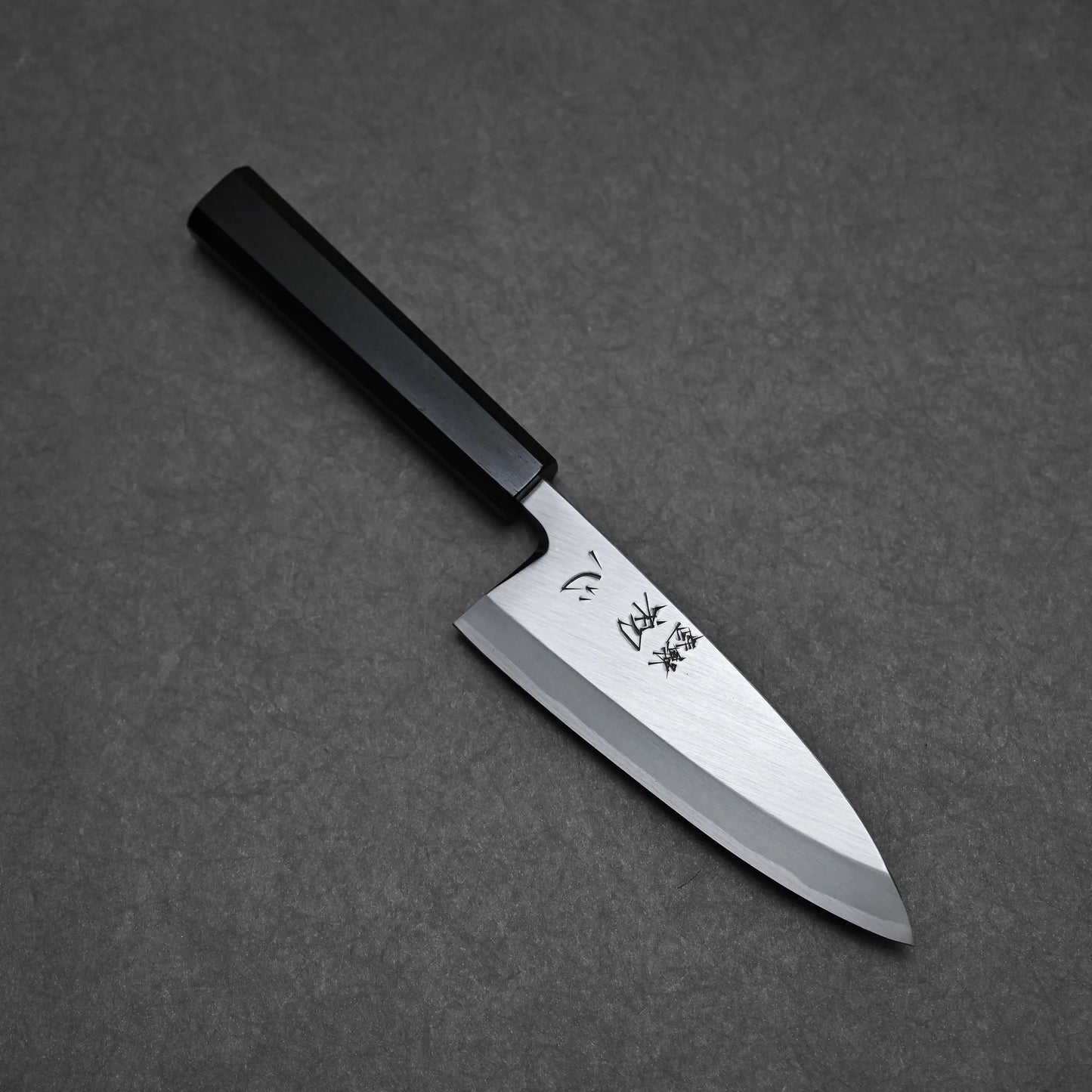 A Hatsukokoro Shirasagi shirogami#2 deba 165mm knife with a black handle and engraved shiny carbon steel blade rests on a dark textured surface.