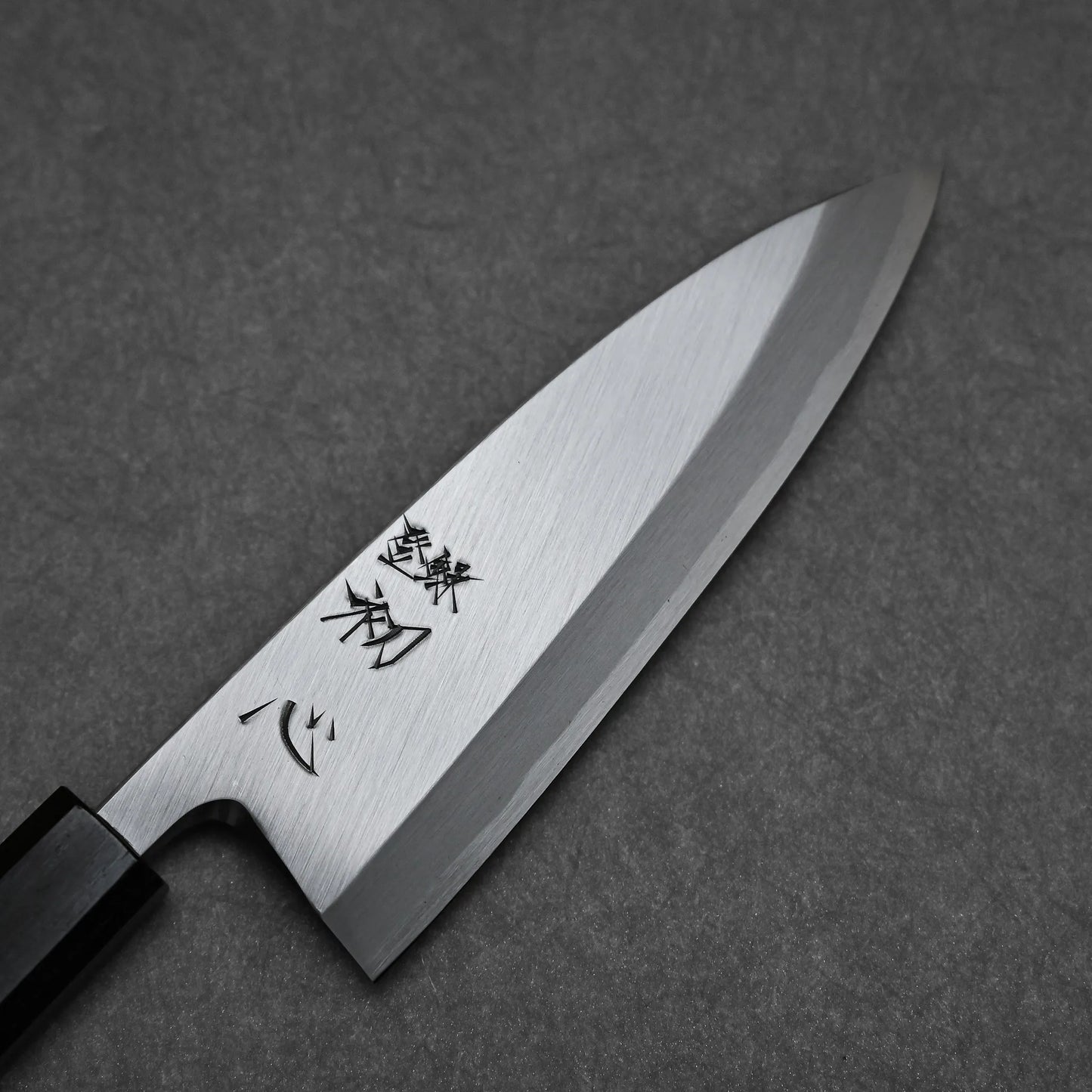 A close-up of the Hatsukokoro Shirasagi Shirogami#2 Deba 165mm, a Japanese fish knife with a black handle and engraved blade, displayed on a dark textured surface.