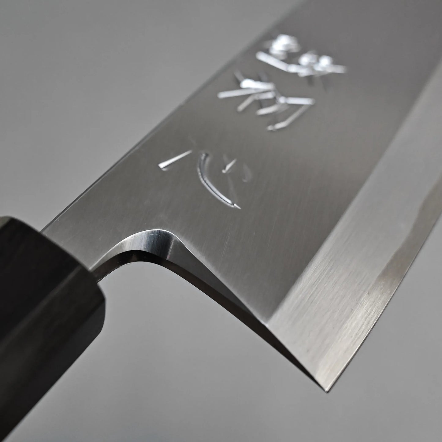 Close-up of the Hatsukokoro Shirasagi shirogami#2 deba 165mm, a Japanese fish knife made from Shirogami#2 carbon steel, displays engraved characters on its sharp blade and a dark handle against a plain grey background.