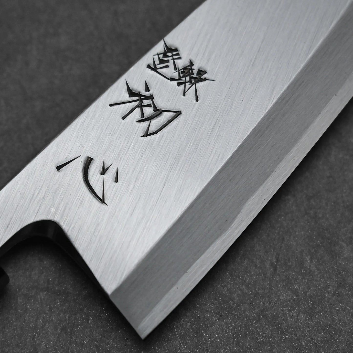 Close-up of the Hatsukokoro Shirasagi shirogami#2 deba 165mm Japanese fish knife, featuring a Shirogami#2 carbon steel blade engraved with Japanese characters, resting on a dark gray surface.