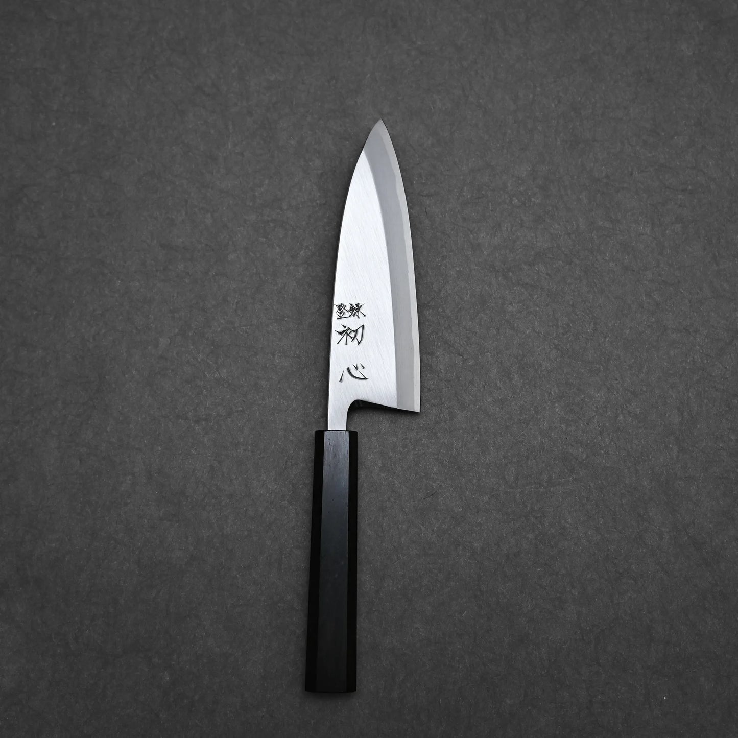 The Hatsukokoro Shirasagi shirogami#2 deba 165mm features a shiny carbon steel blade with engraved characters and a black handle, shown on a dark gray textured surface.
