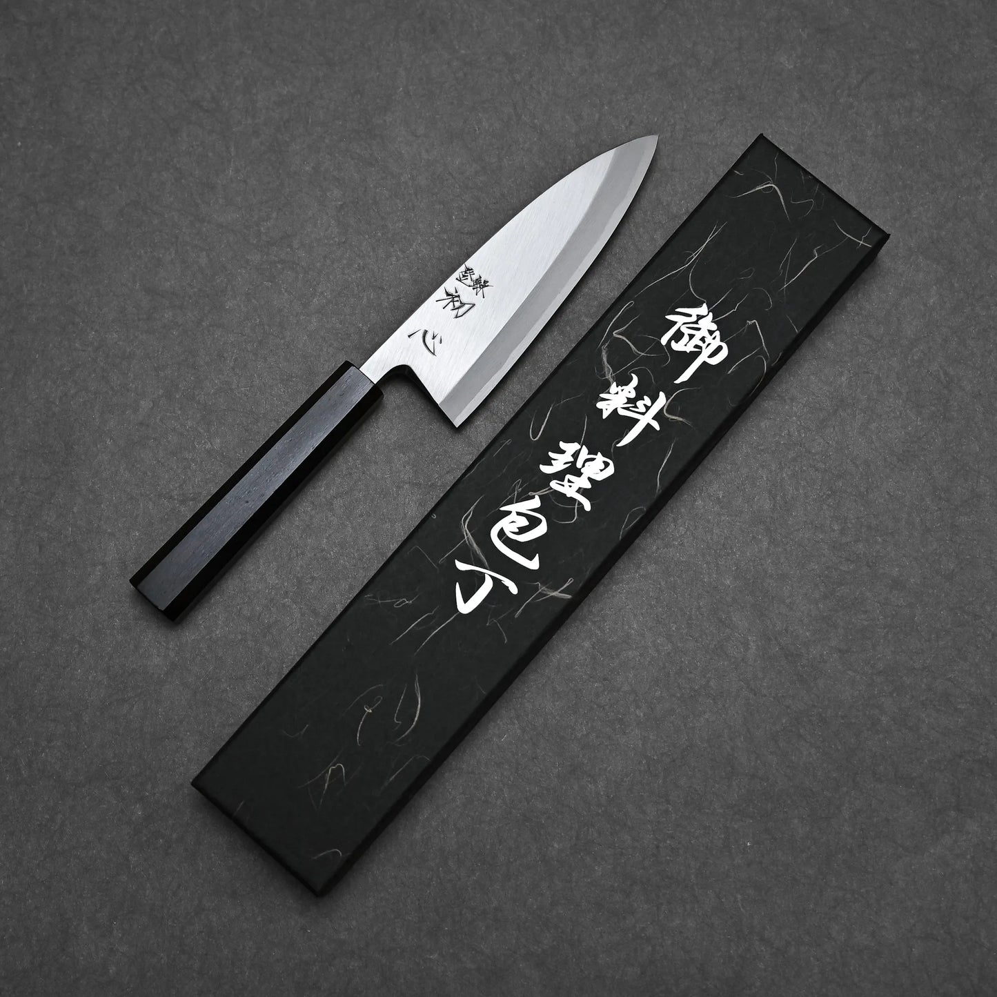 The Hatsukokoro Shirasagi Shirogami#2 Deba 165mm, featuring a black handle, lies beside a stylish black box with white Japanese characters on a dark textured surface, showcasing traditional craftsmanship and precise cutting performance.