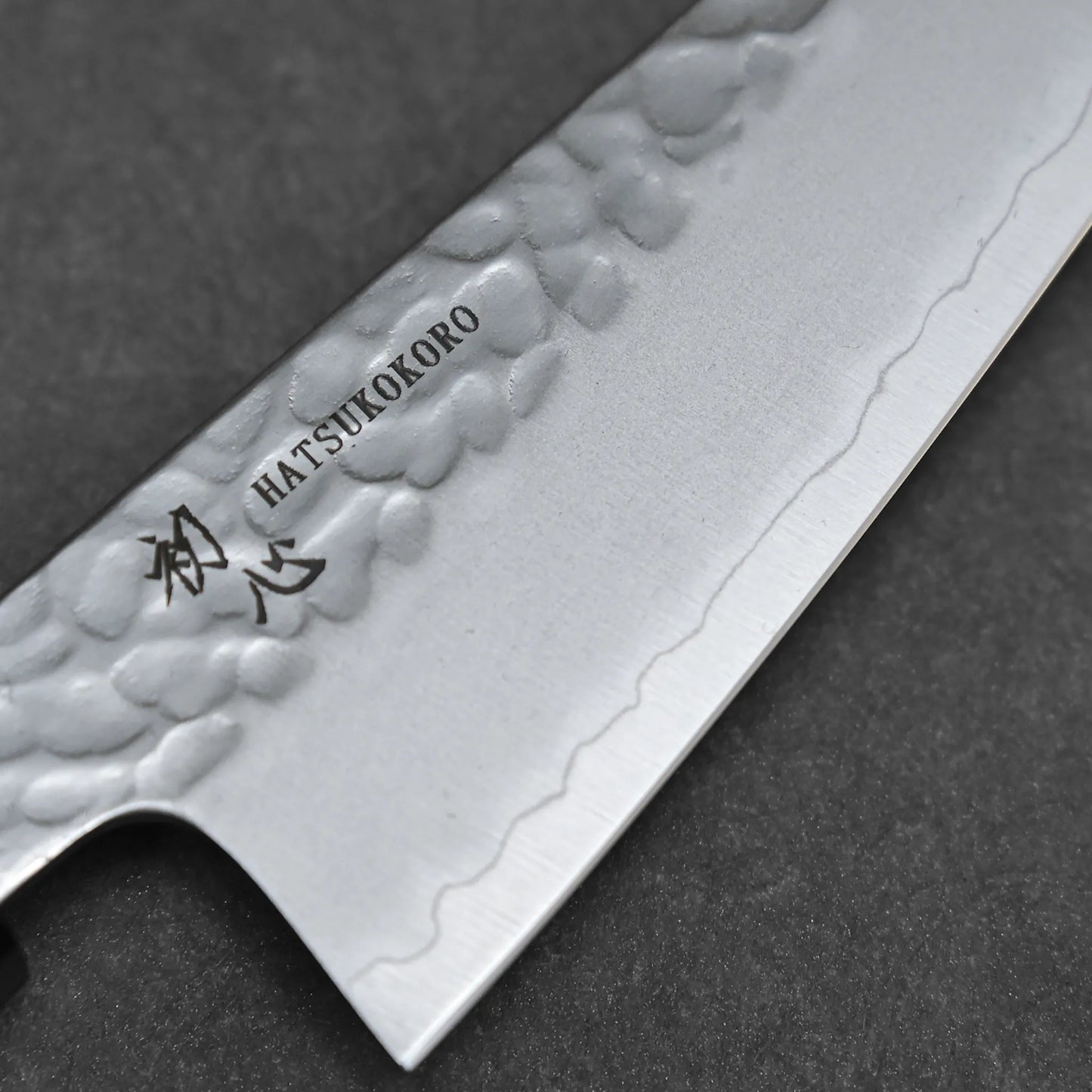 Close-up of the 150mm Hatsukokoro tsuchime ginsan honesuki, showcasing its hammered finish and engraved Japanese characters on the blade, set against a dark background.