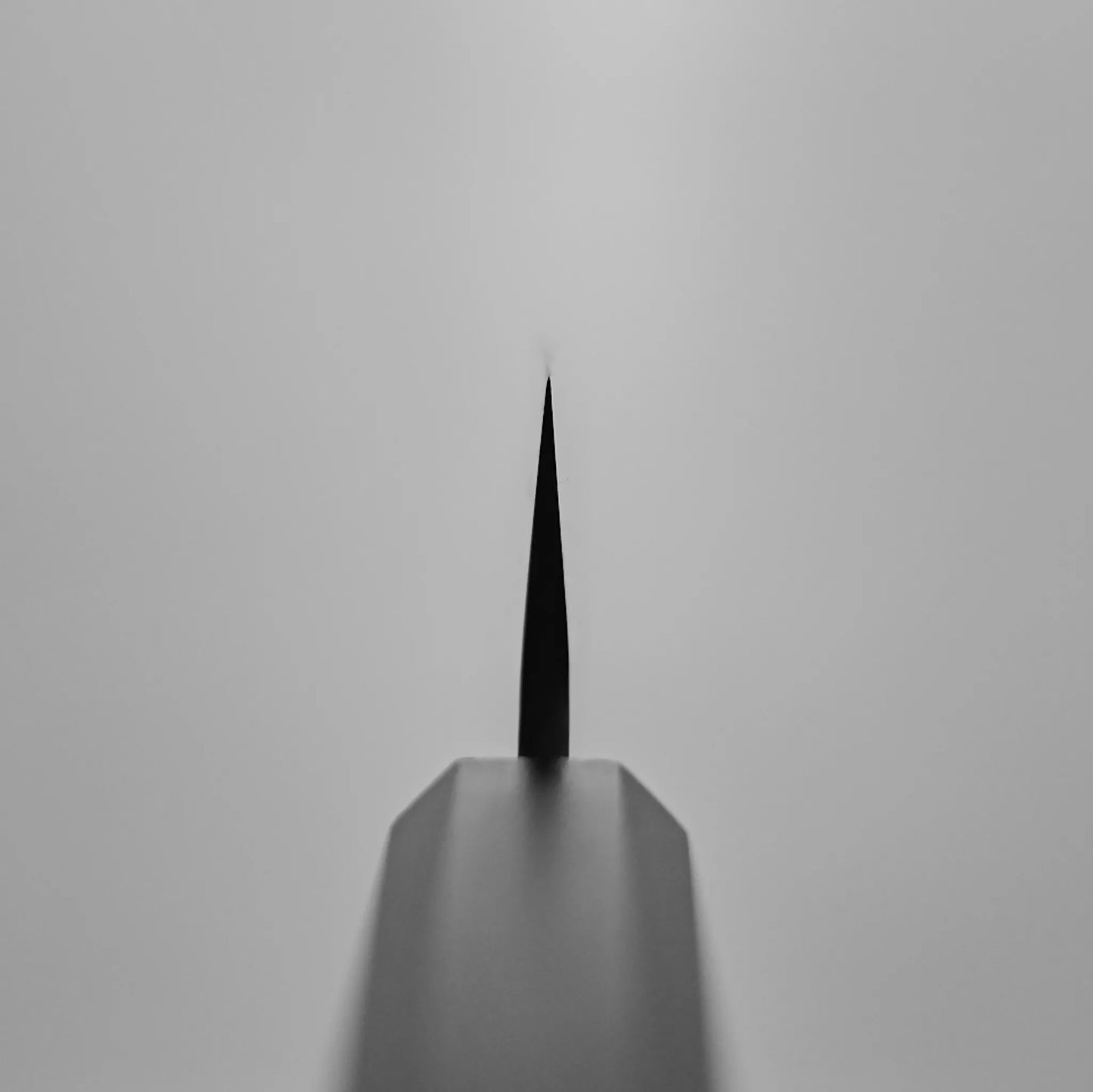 Close-up, black-and-white photo of the pointed tip of a Hatsukokoro tsuchime ginsan honesuki 150mm, shot from the handle end with sharp focus on the knife’s edge and a blurred background.