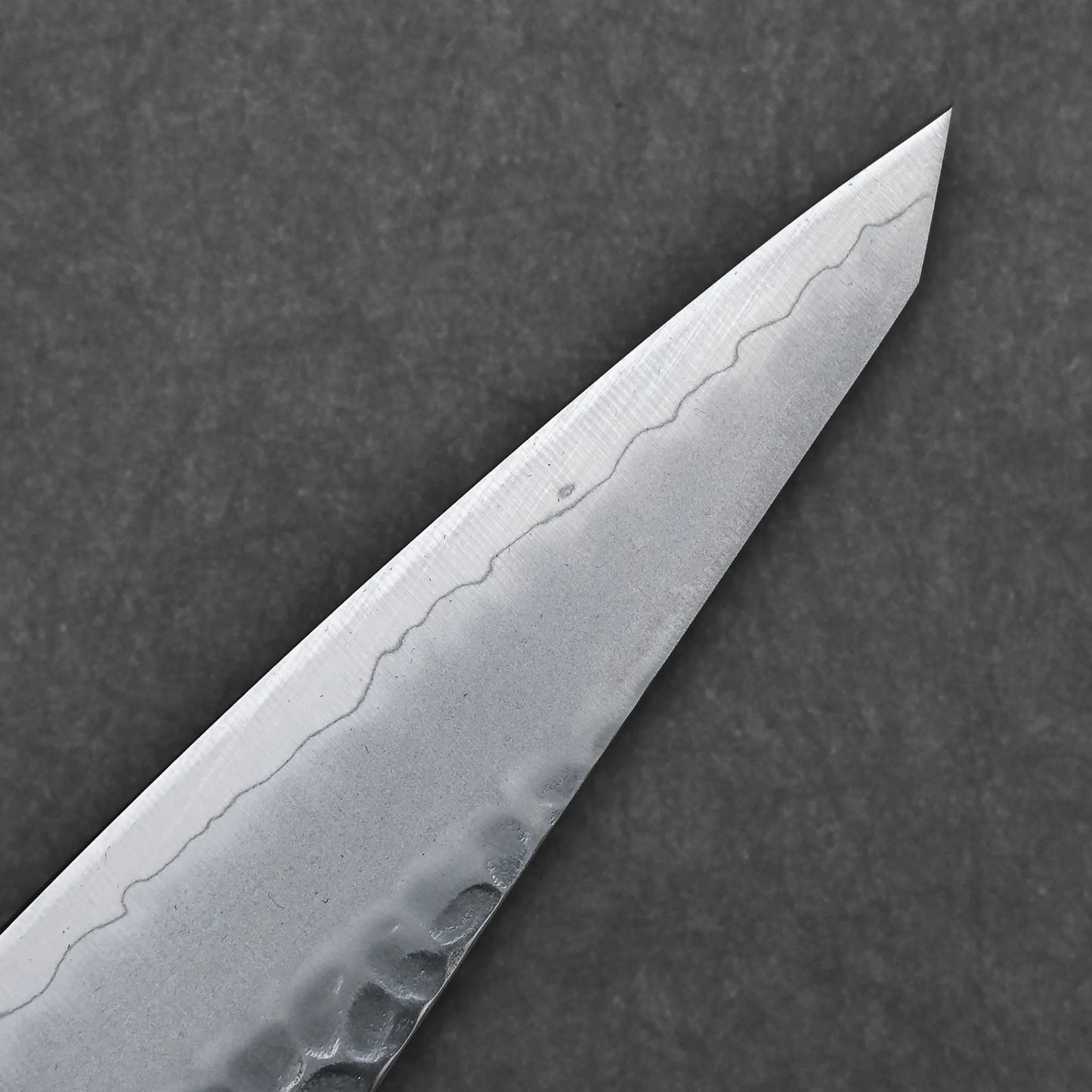 Close-up of the Hatsukokoro tsuchime ginsan honesuki 150mm knife tip, showcasing its hammered tsuchime finish and wavy blade pattern, set against a dark, textured background.