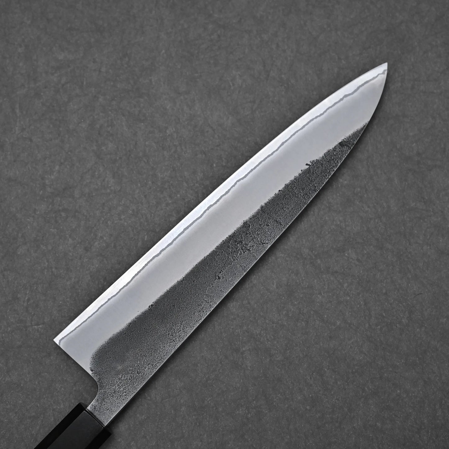A close-up of the Hatsukokoro x Yoshikane kurouchi nashiji shirogami#2 gyuto 210mm highlights its sharp, layered blade and textured finish against a dark gray textured background.