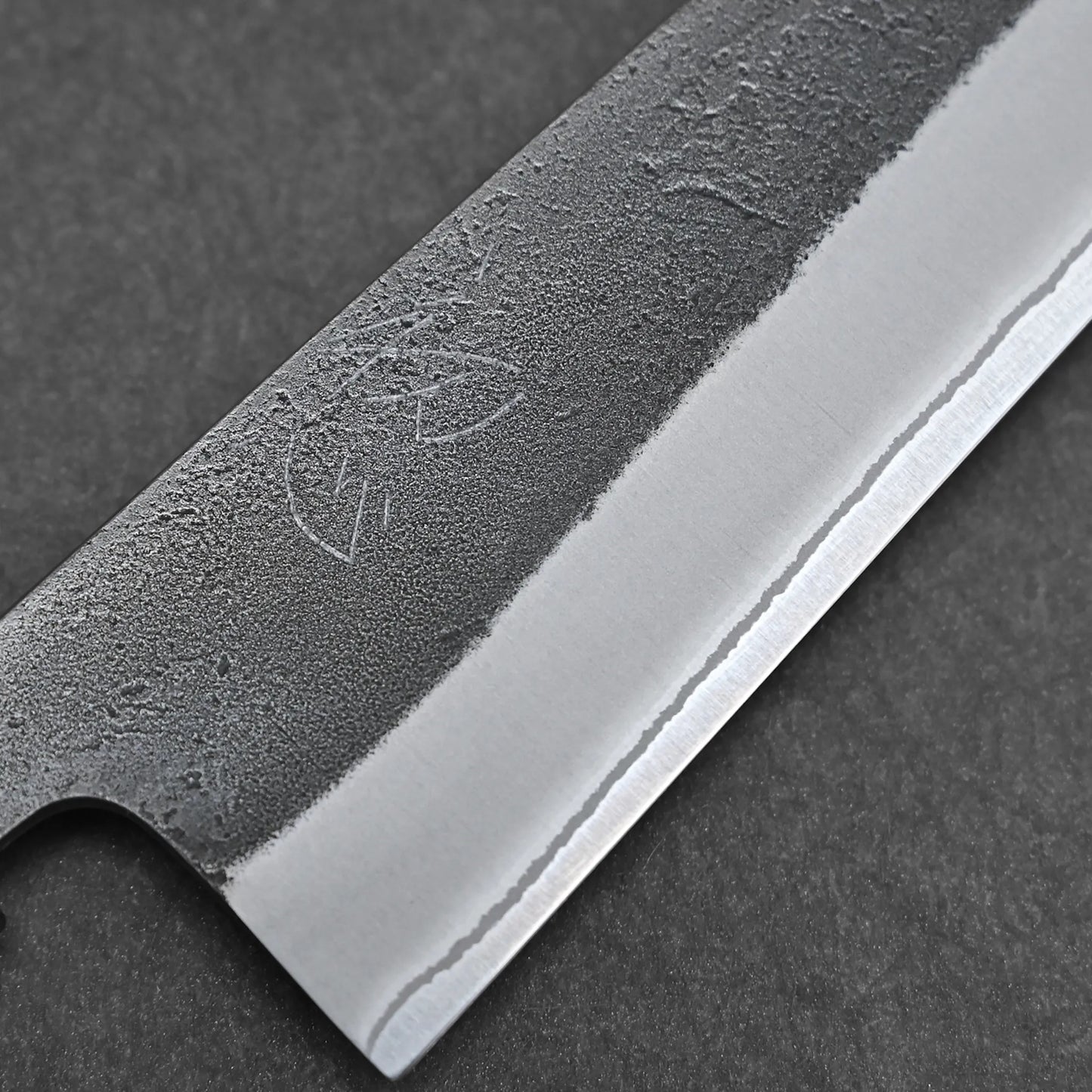 Close-up of a Hatsukokoro x Yoshikane kurouchi nashiji shirogami#2 gyuto 210mm blade, showing its textured matte finish and polished edge with faint etching against a dark background.