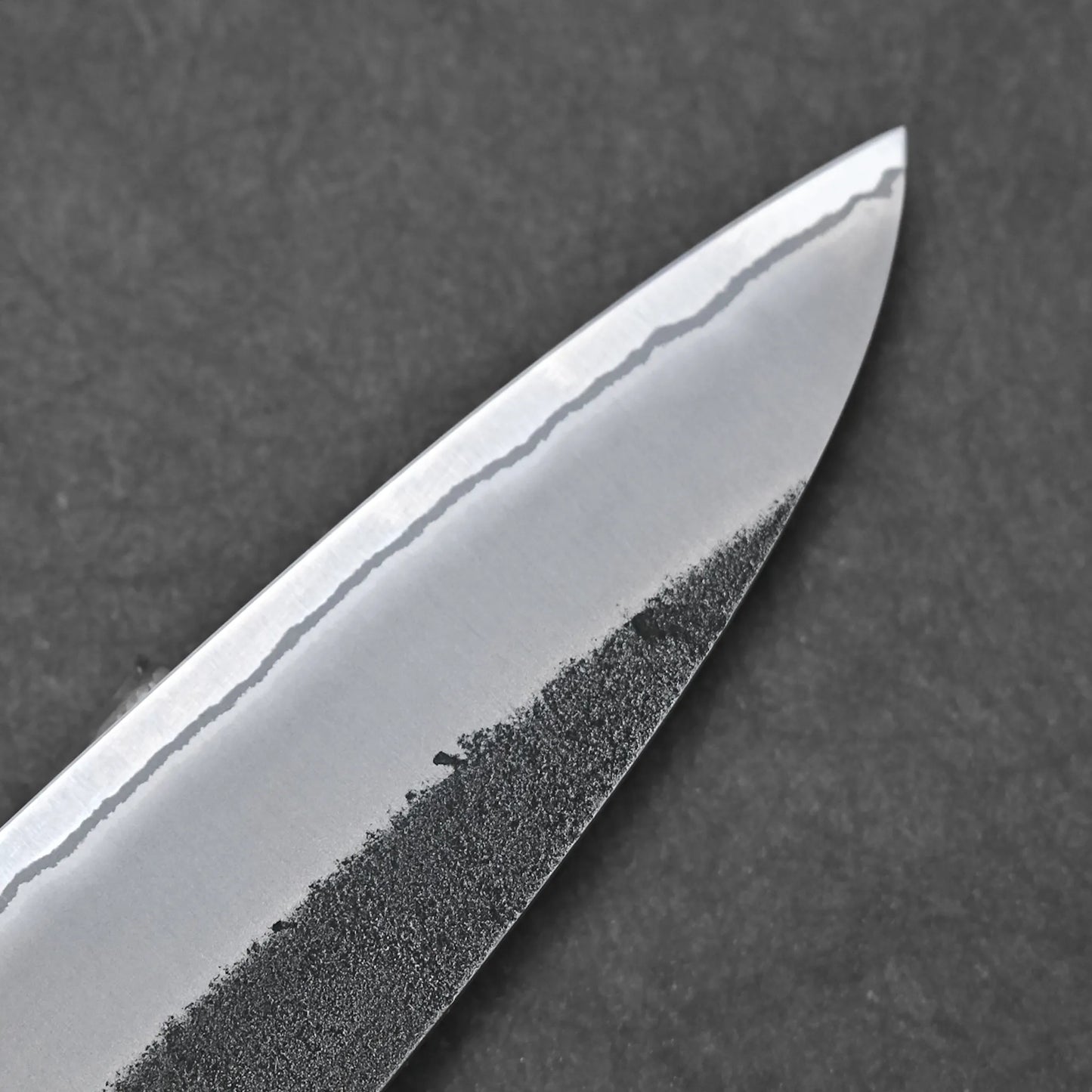 Close-up of the Hatsukokoro x Yoshikane kurouchi nashiji shirogami#2 gyuto 210mm tip, showing its wavy hamon and textured dark lower blade against a dark background.