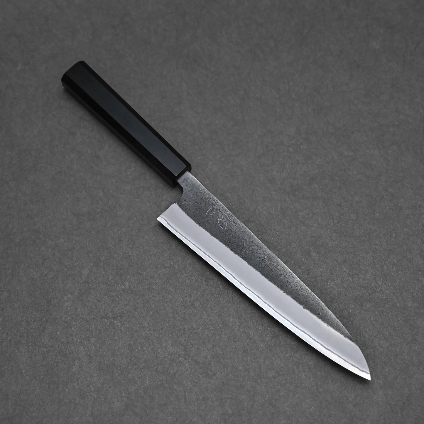The Hatsukokoro x Yoshikane kurouchi nashiji shirogami#2 gyuto 210mm, featuring a rectangular black handle and a shiny pointed blade, rests on a dark gray textured surface.