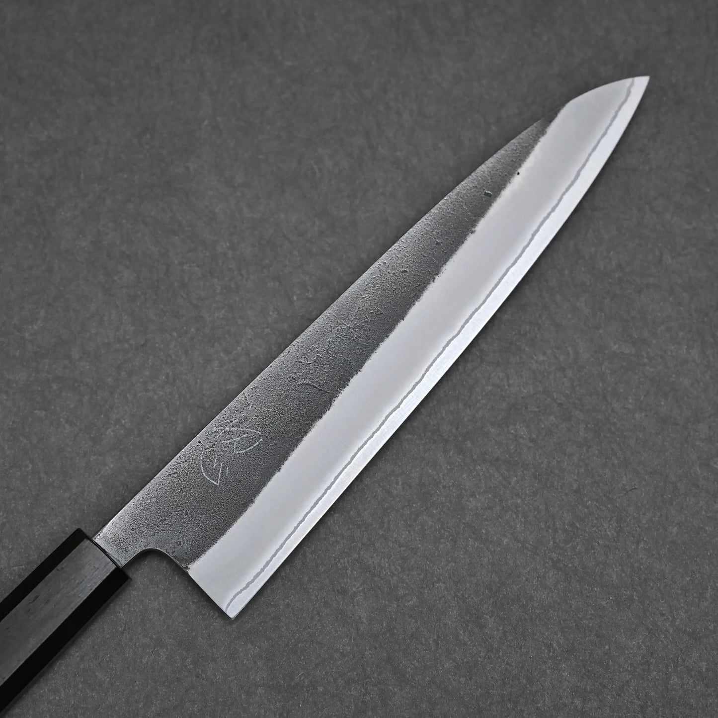 A close-up of the Hatsukokoro x Yoshikane kurouchi nashiji shirogami#2 gyuto 210mm, featuring a sharp pointed blade and dark handle, crafted from Shirogami#2 carbon steel, displayed on a gray textured surface.