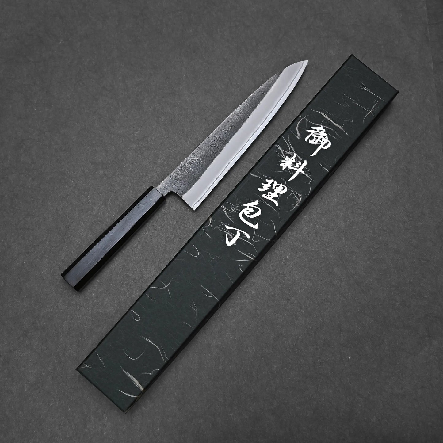 A Hatsukokoro x Yoshikane kurouchi nashiji shirogami#2 gyuto 210mm with a black handle sits next to its matching box with white Japanese characters, set against a dark backdrop. The blade is crafted from premium Shirogami#2 carbon steel.