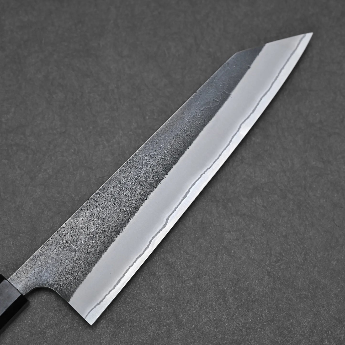 A close-up of the Hatsukokoro x Yoshikane kurouchi nashiji shirogami#2 kiritsuke gyuto 210mm, featuring a sharp pointed blade with a wavy pattern, set against a dark gray textured background.