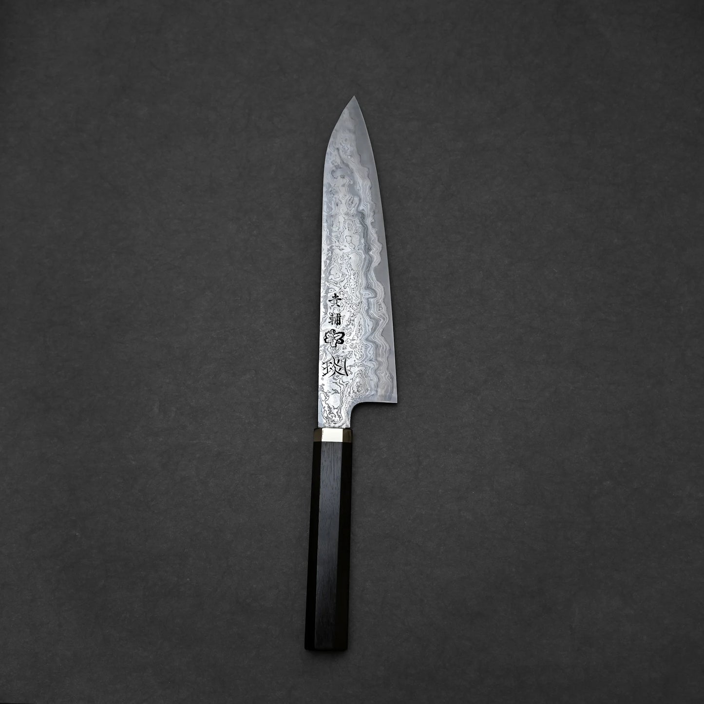 Japanese chef’s knife with Damascus steel blade featuring swirling layered forge patterns and vertically engraved kanji characters; dark wooden handle with metal bolster, photographed on textured surface.