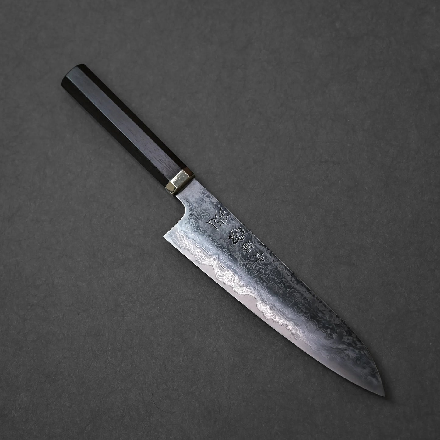 Japanese chef’s knife with Damascus steel blade featuring intricate layered forge patterns, vertically engraved kanji characters, and a dark wooden handle with brass bolster; photographed against a textured backdrop.