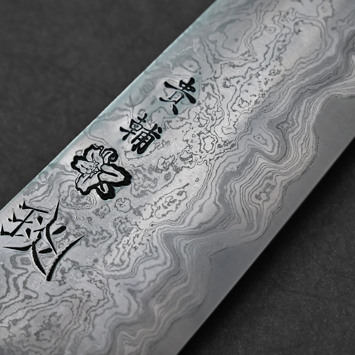 Japanese Damascus steel knife blade featuring layered wavy forge patterns, vertically engraved kanji characters, and a delicate flower motif etched near the center.