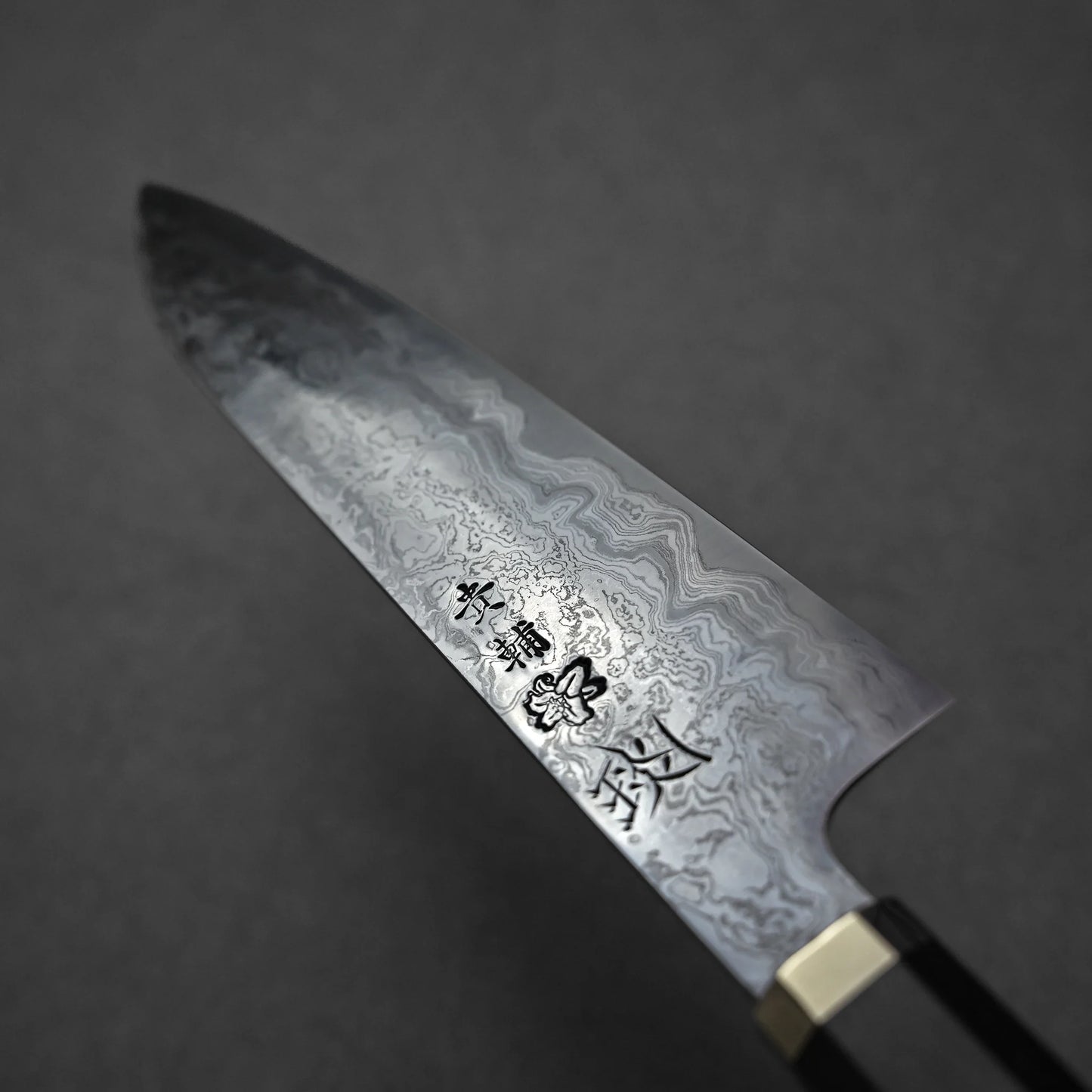Close-up of a Japanese Damascus steel knife blade with intricate layered forge patterns and vertically engraved kanji characters; photographed against a dark background.