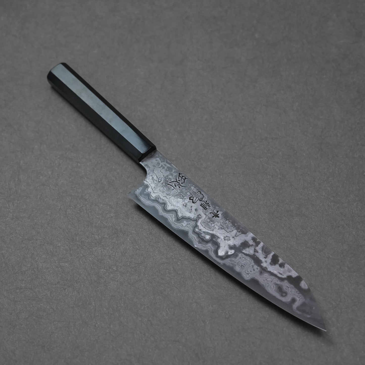 Japanese chef’s knife with Damascus steel blade, featuring finely layered forge patterns, dark octagonal handle, and engraved kanji near the base.
