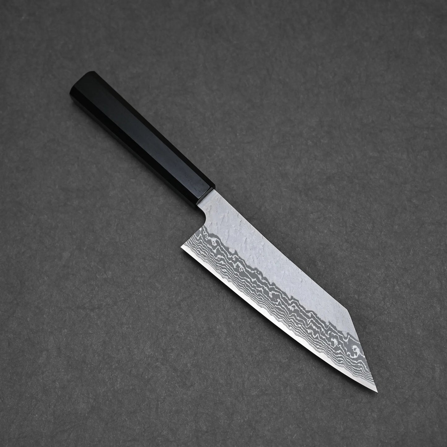 The Nigara tsuchime damascus VG10 bunka 180mm is a Japanese-style kitchen knife with a patterned, angular Takefu VG10 stainless steel blade and black handle, elegantly displayed on a dark gray surface.