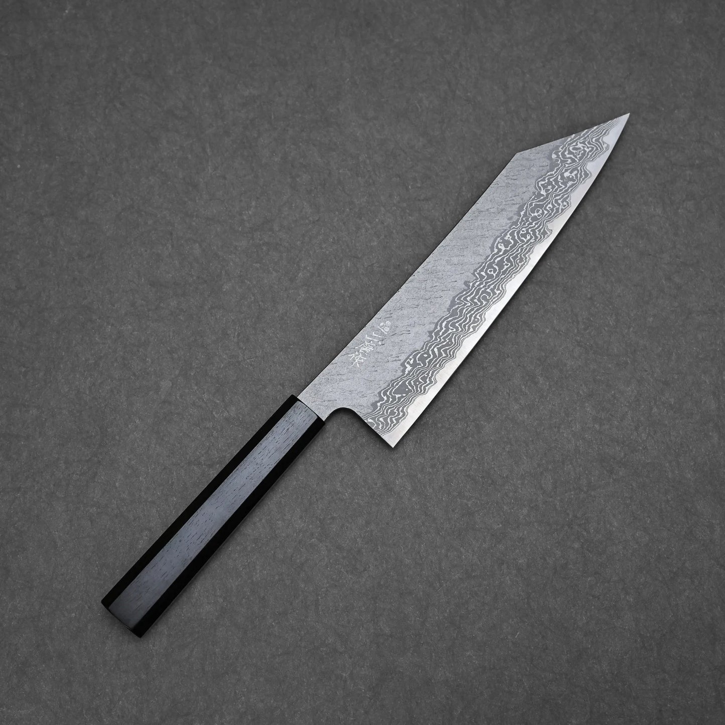 A Nigara tsuchime damascus VG10 kiritsuke gyuto 210mm kitchen knife with a black handle and patterned steel blade, photographed on a dark gray textured surface. The blade features distinctive wavy Damascus patterns.