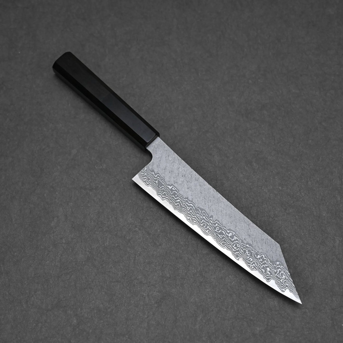 The Nigara tsuchime damascus VG10 kiritsuke gyuto 210mm, featuring a black handle and patterned steel blade, is set on a dark gray surface.