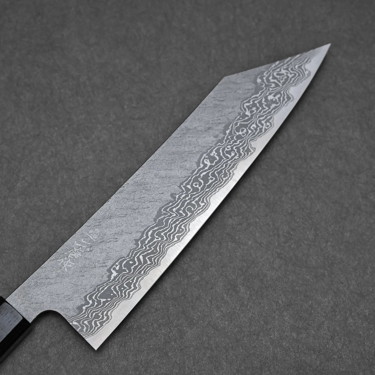 A close-up of the Nigara tsuchime damascus VG10 kiritsuke gyuto 210mm shows its wavy, layered blade and dark handle, set against a dark, textured background.