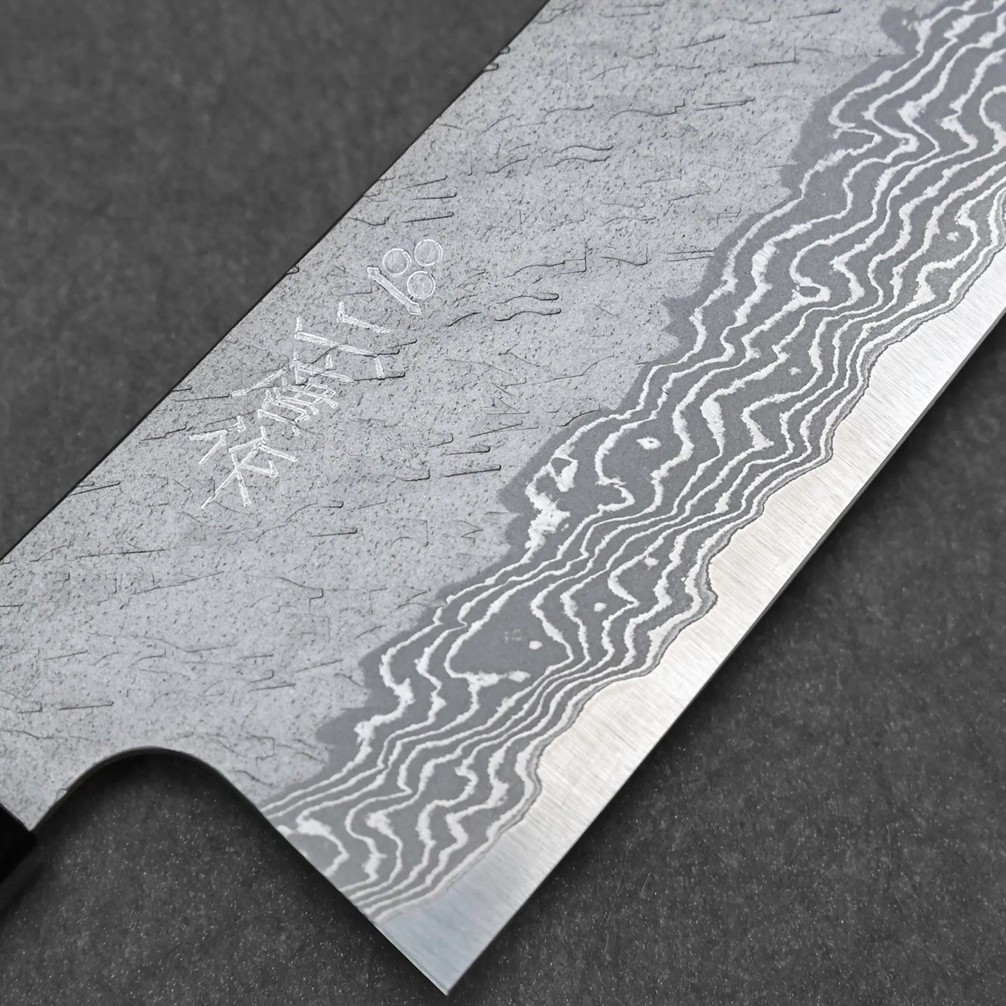 Close-up of the Nigara tsuchime damascus VG10 kiritsuke gyuto 210mm blade, showing intricate patterns, a sharp edge, and engraved markings against a dark background.