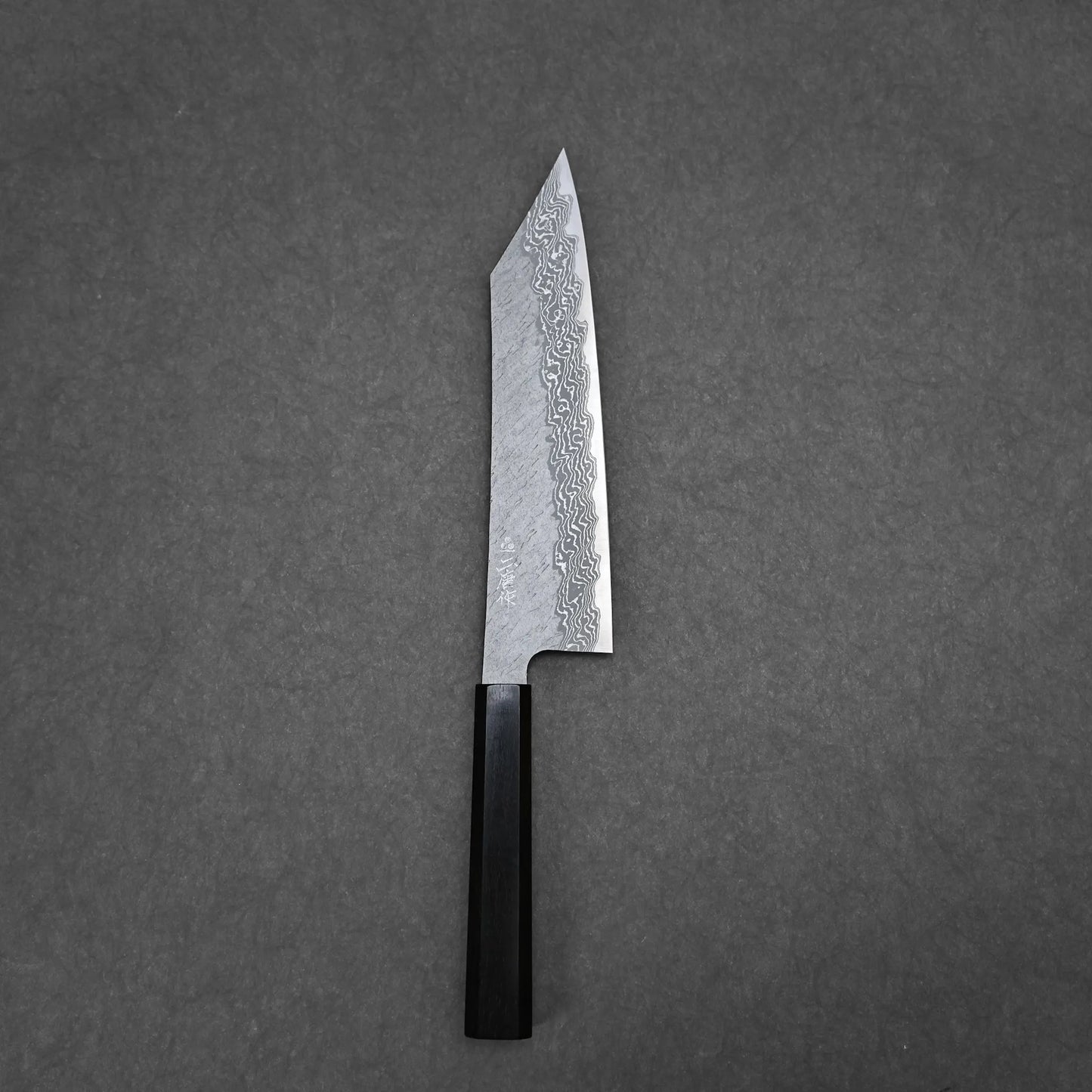 The Nigara tsuchime damascus VG10 kiritsuke gyuto 210mm, featuring a dark wooden handle, is displayed on a plain gray surface.